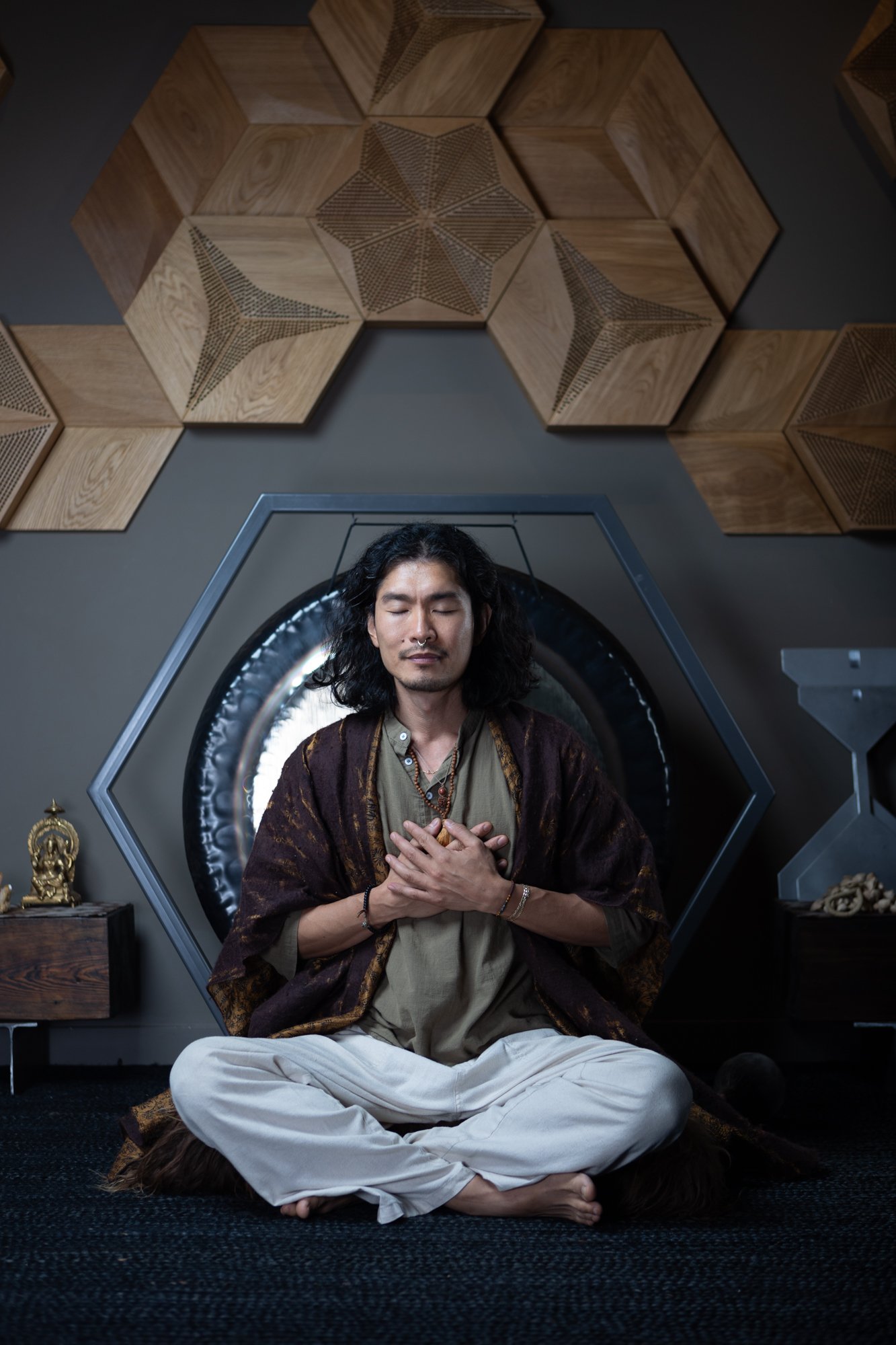 A man with long curly hair and closed eyes, sitting cross-legged with hands on chest, in front of a metallic circular structure. The background features wooden hexagonal panels with intricate geometric designs.
