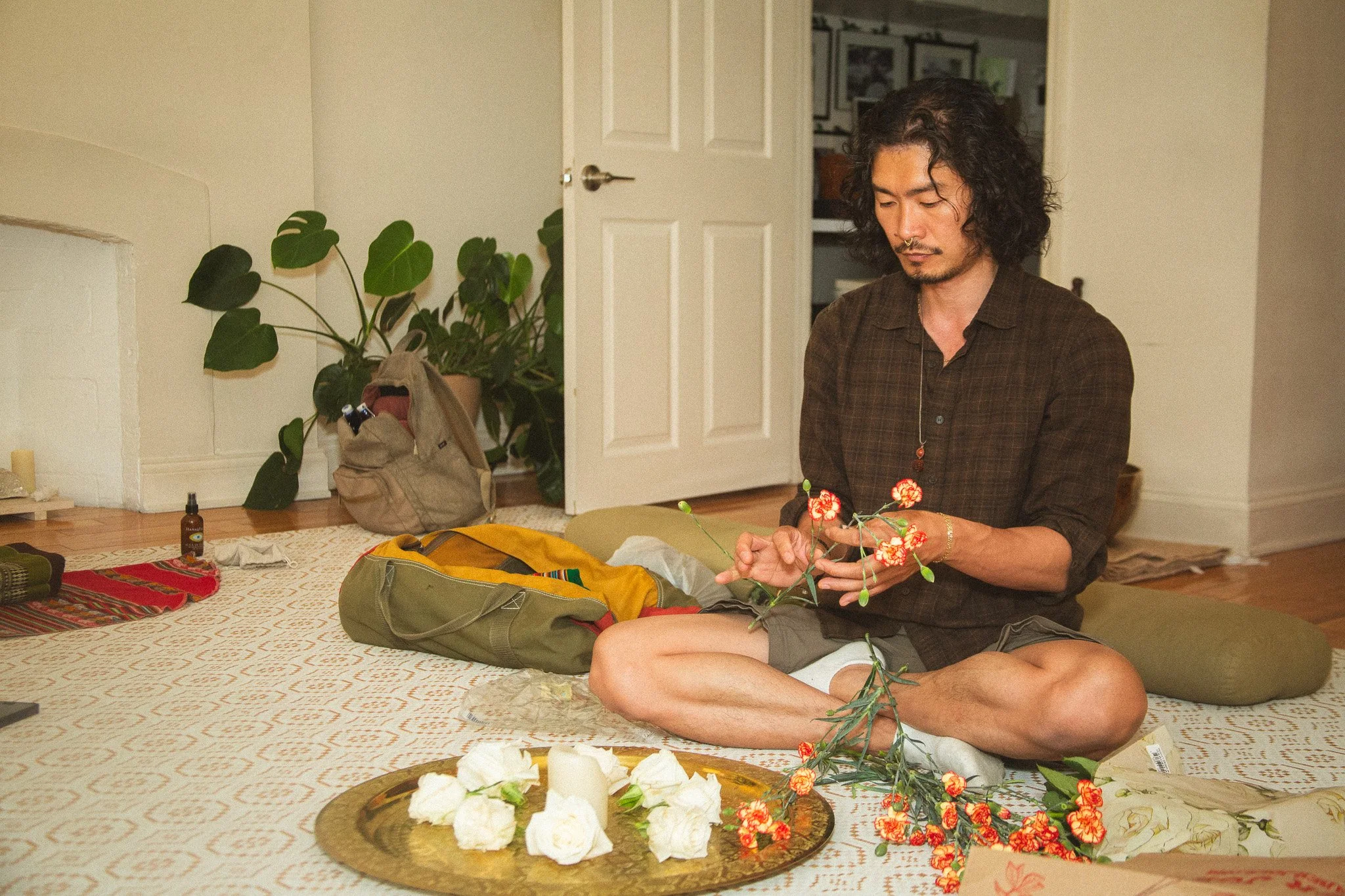 A man with long curly black hair and a mustache seated on a rug, holding a flowered branch, in a room decorated with plants, candles, and various items.
