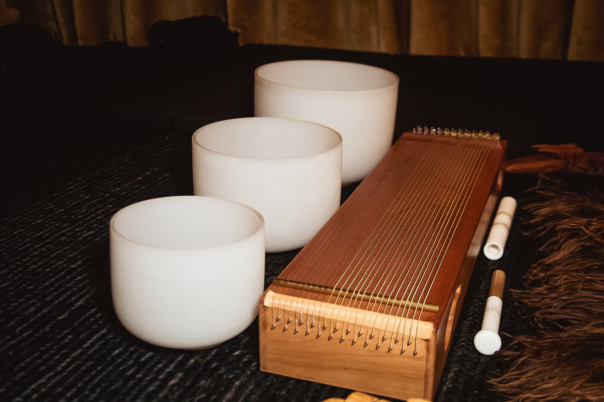 Three white singing bowls, a wooden kalimba, and a mallet placed on a dark textured surface with a wooden wall background.