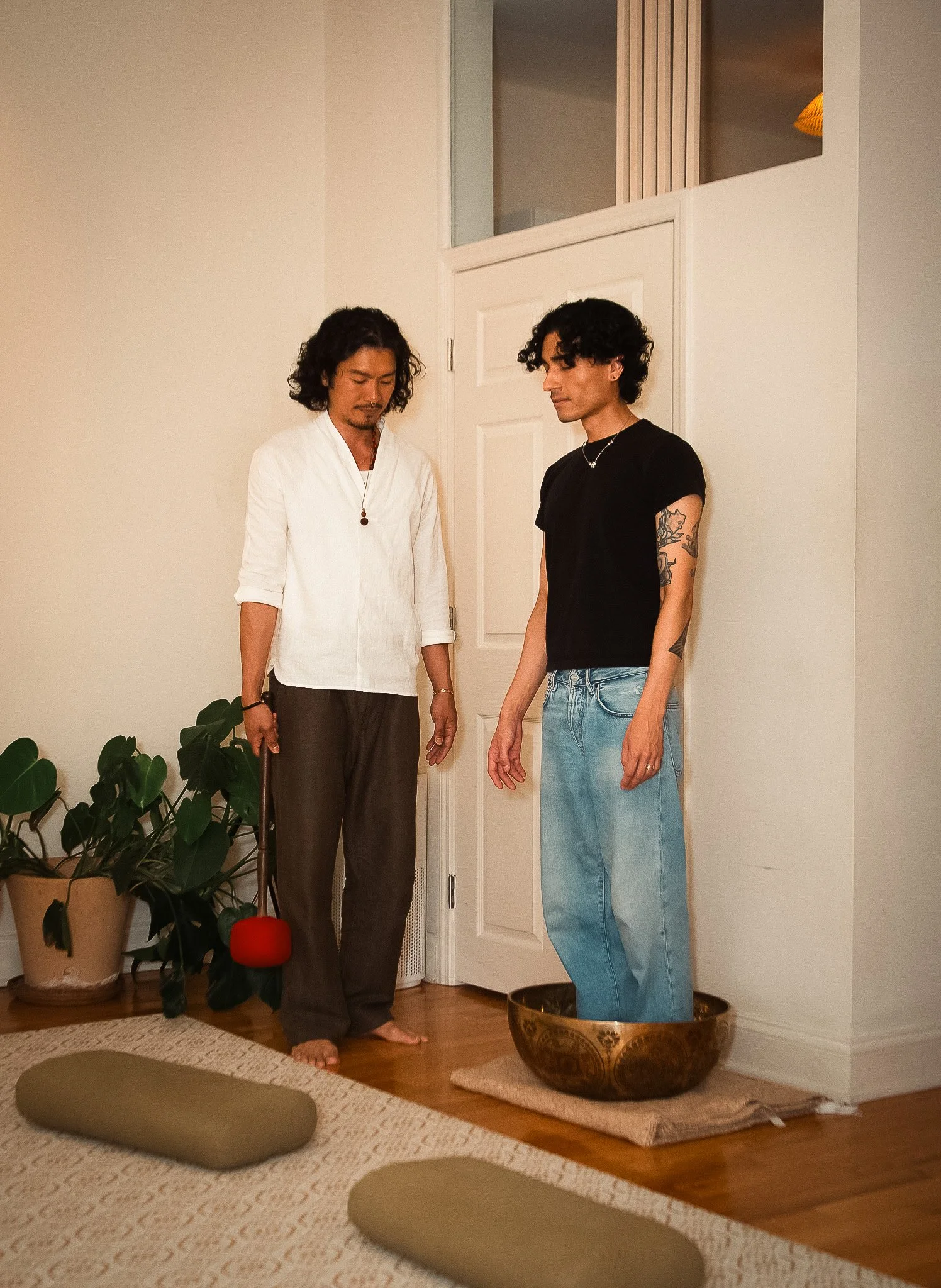 Two men standing in a room, one with long curly dark hair and wearing a white top, the other with short curly dark hair and sitting in a large bowl, both with their eyes closed and head tilted downward. The room has white walls, a small door, a potted plant, and a meditation cushion on the floor in the foreground.