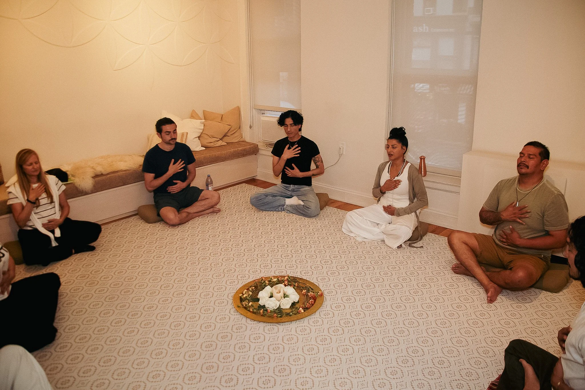 A group of people practicing meditation, sitting cross-legged on the floor with their eyes closed and hands placed over their hearts, in a softly lit room with minimal decor and a large decorative tray with flowers in the center.