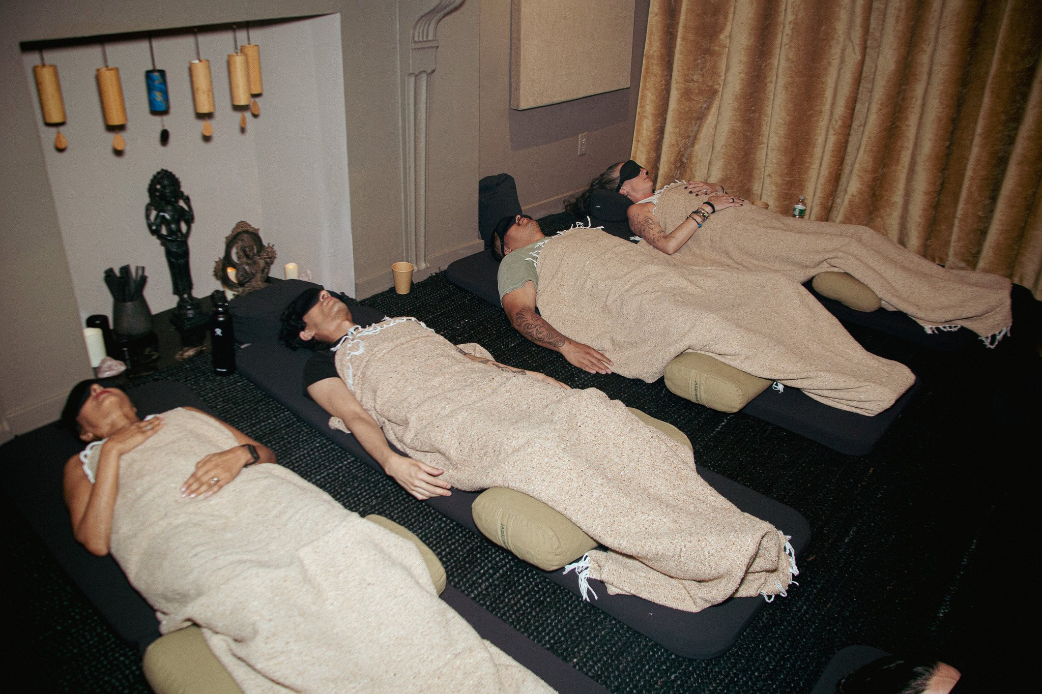 Four people are lying on massage tables with beige blankets, wearing eye masks, in a room with beige curtains and Buddhist statues and decorations on the wall.