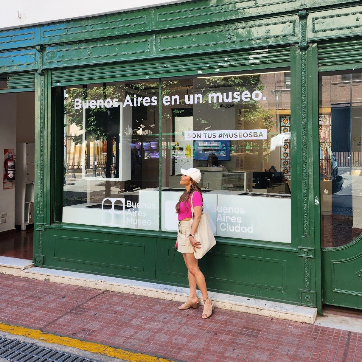 A woman in a pink top, beige shorts, and beige heels stands outside the Buenos Aires Museo. She wears a white hat and carries a beige tote bag. The museum has a large green storefront window with white text that reads "Buenos Aires en un museo" and signing that says "Son tus #MuseosBA". The street in front has red brick pavement and a yellow line.