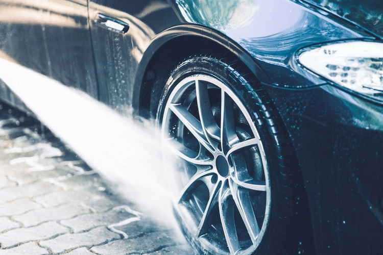 A close-up of a black car being washed with a stream of water from a hose, focusing on the wheel and tire.