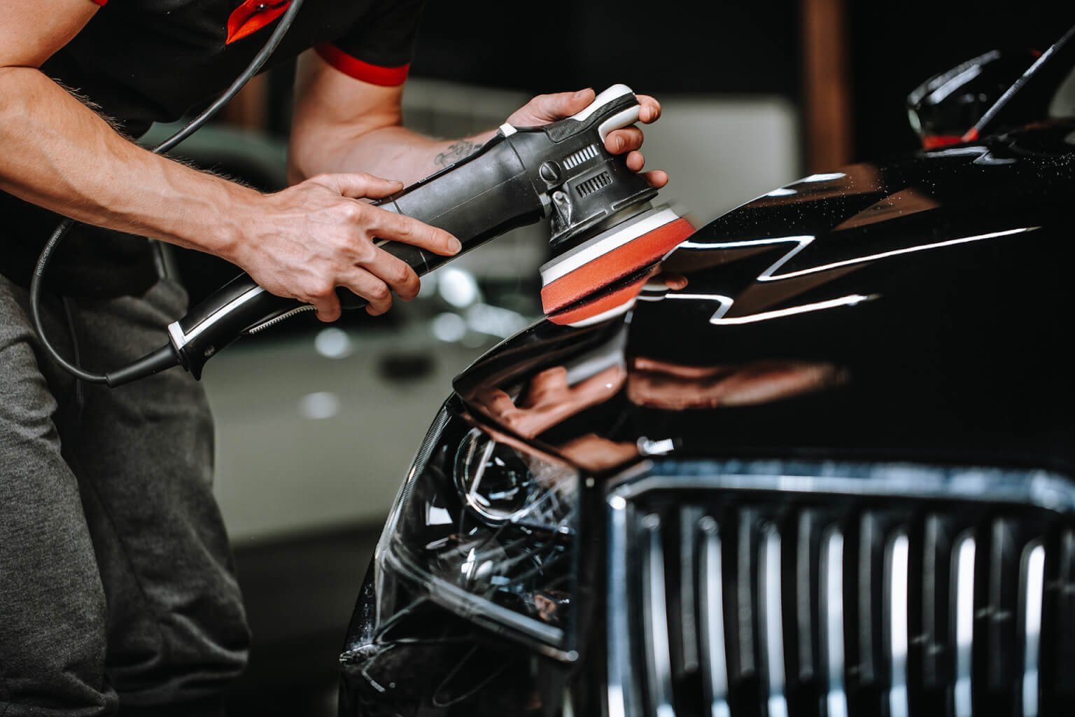 Person polishing a black car with a handheld electric buffer and foam pad.