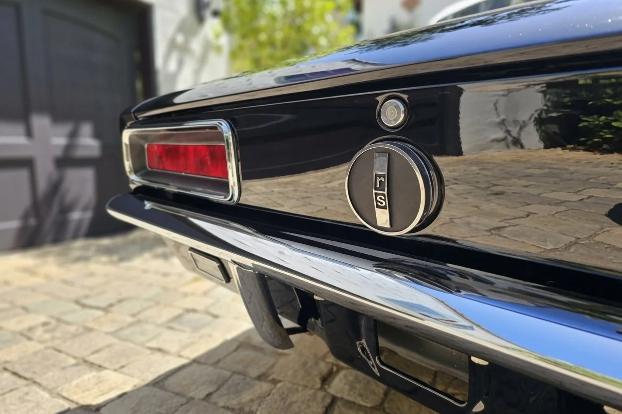 Close-up of the rear end of a classic black car, showing the tail light, a parking sensor, and an 'RS' badge on the trunk lid, with a cobblestone driveway background.