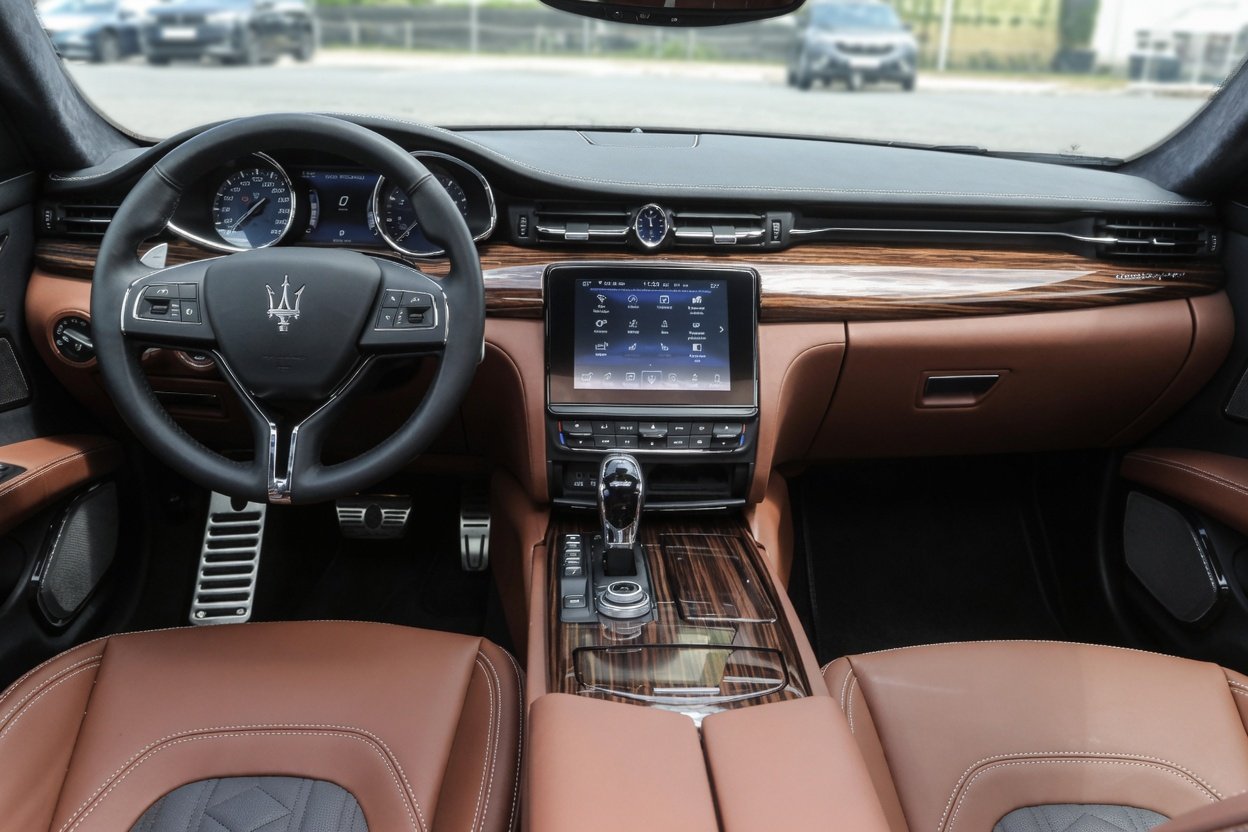 Interior of a luxury car with a leather dashboard, center console, and steering wheel, featuring a modern touchscreen display and a woodgrain finish.