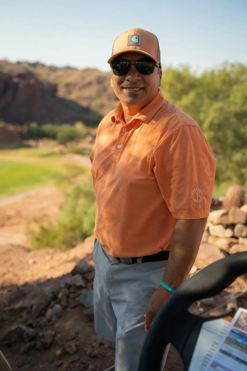 A man wearing sunglasses, a brown hat, an orange polo shirt, and gray pants, standing outdoors with a landscape of hills and trees in the background.