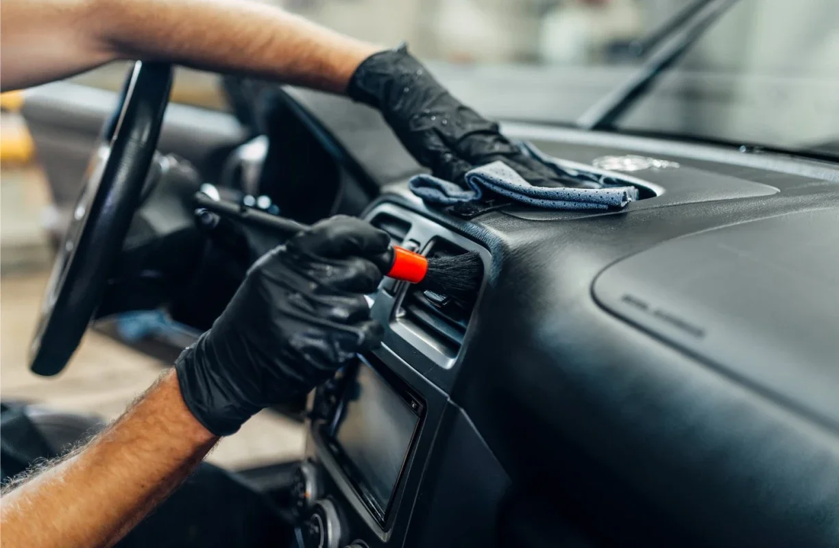 Person wearing black gloves cleaning car air vents with a brush and cloth.
