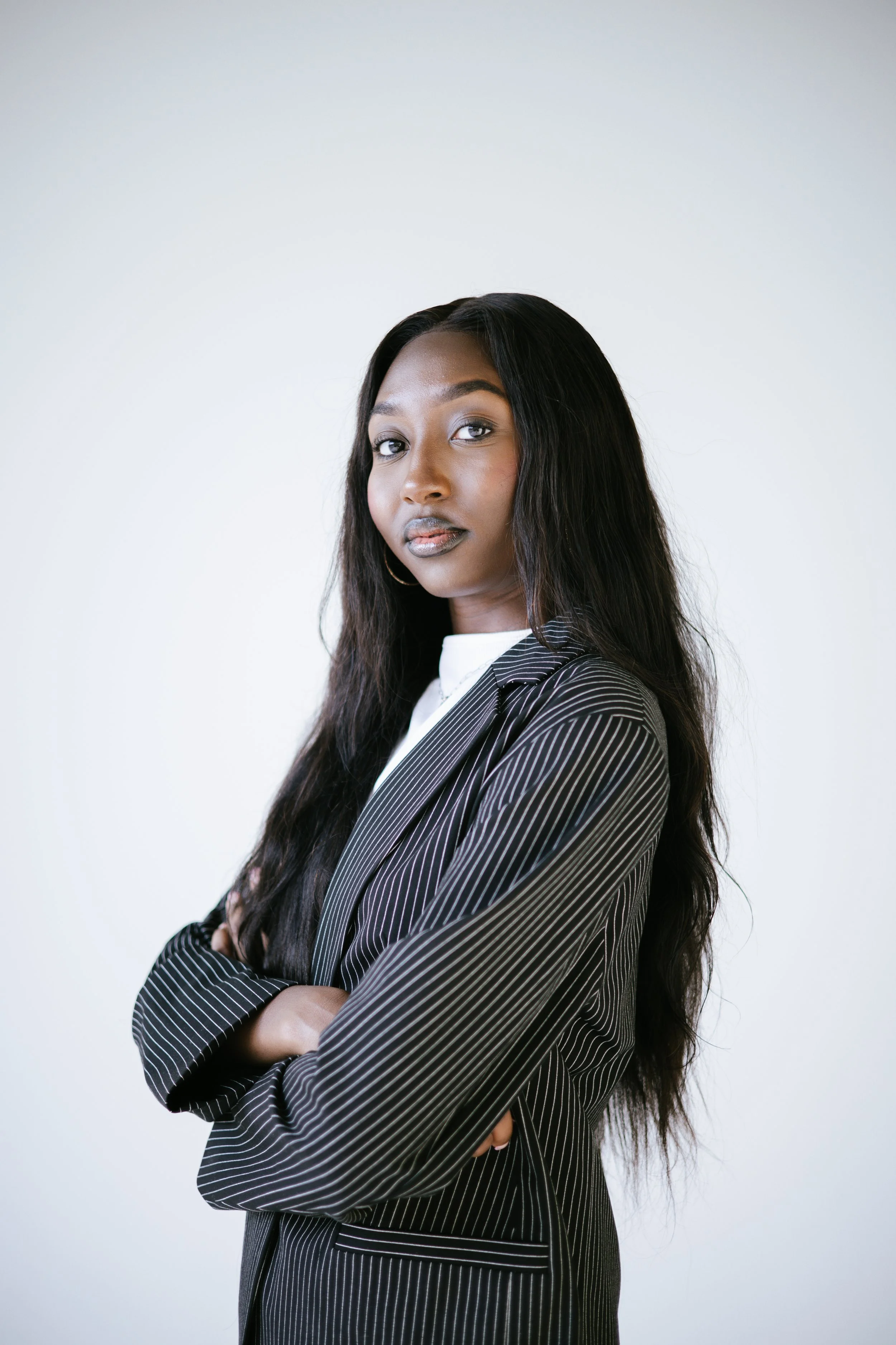A confident Black woman with long black hair, wearing a black and white pinstripe blazer and crossing her arms, standing against a plain white background.