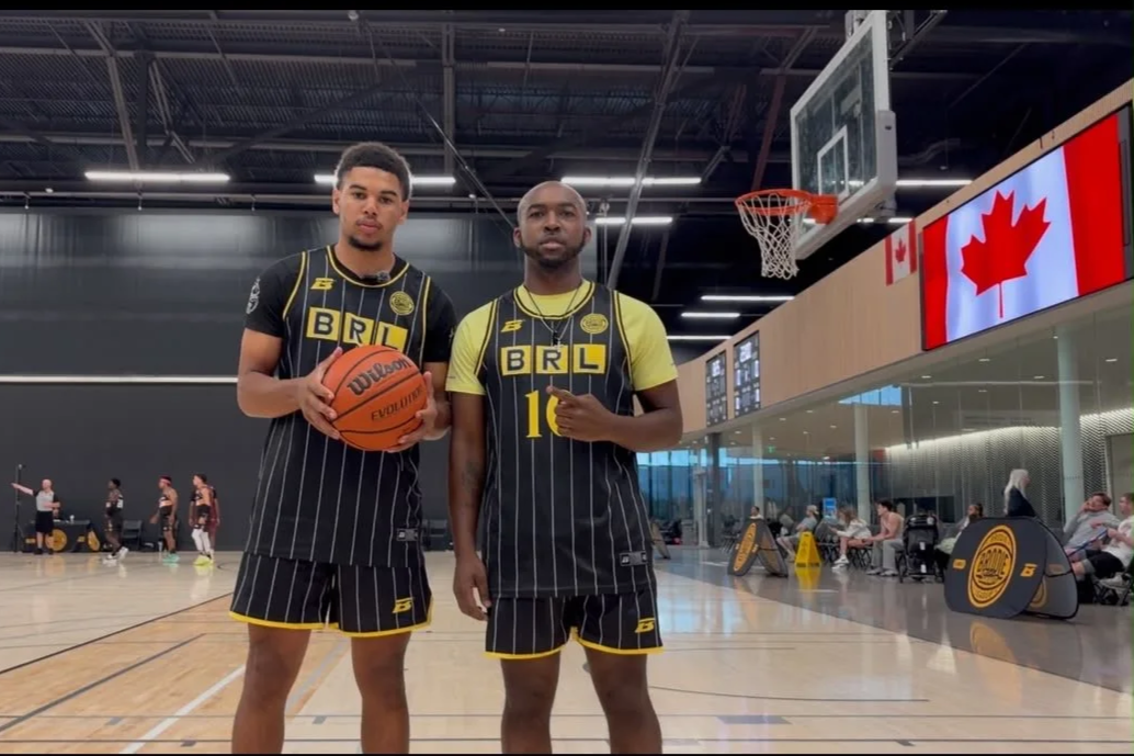 Two basketball players in black and yellow uniforms standing inside a basketball court, with one holding a basketball, and a large Canadian flag displayed on a digital screen above them.