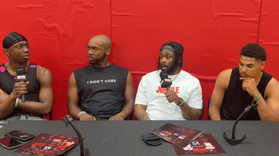 Four men sitting at a table during an interview or panel. The table has posters, microphones, and electronic devices. The background is red.