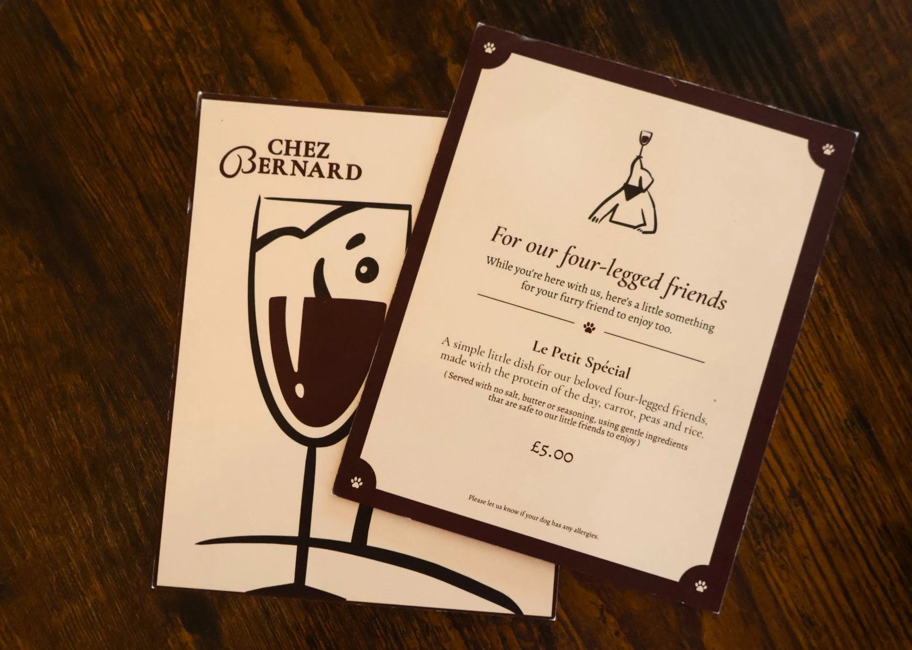 Menu cards on a wooden table, one with a drawing of a glass with a face, and another with text about a dish called 'Le Petit Spécial' for four-legged friends.