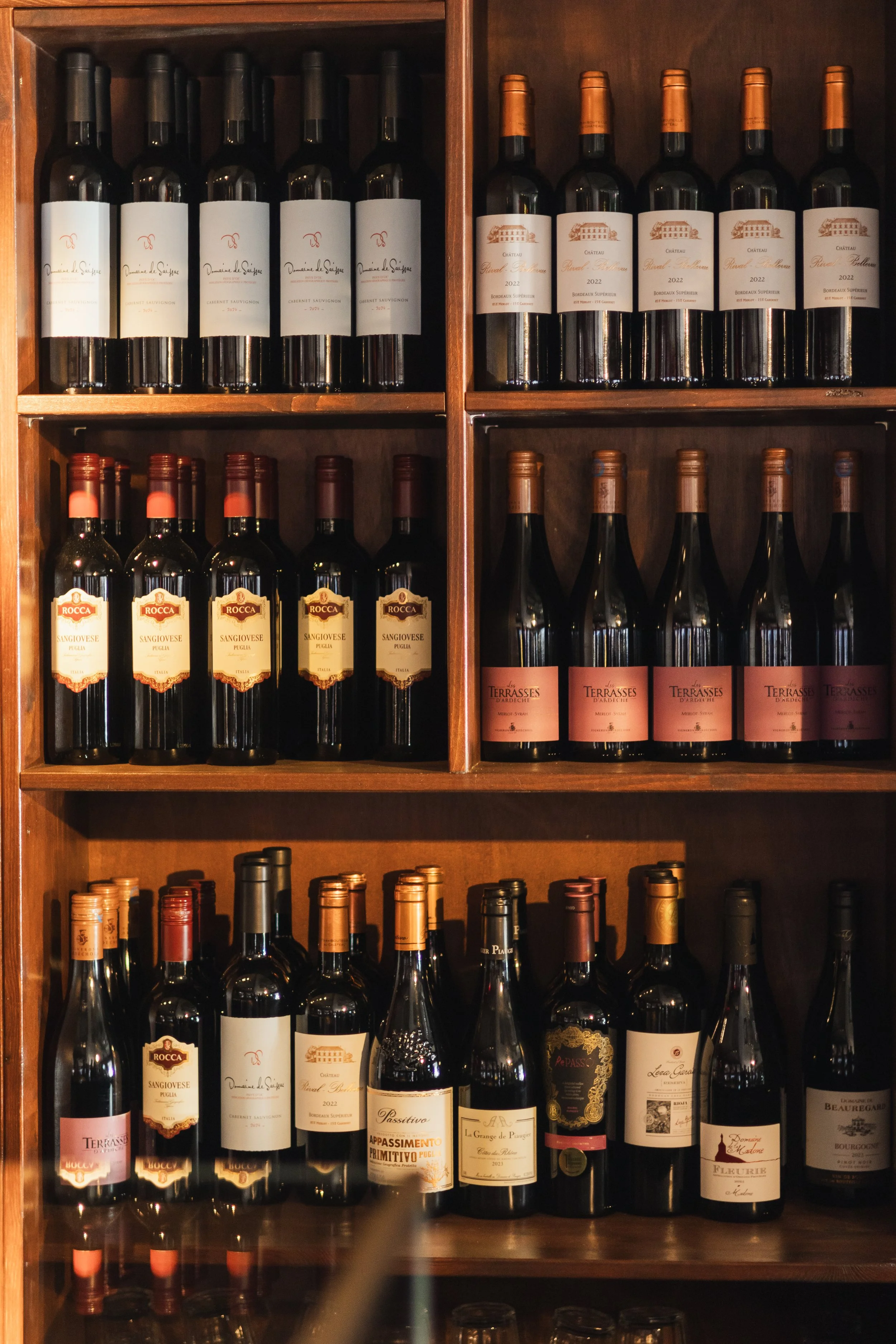 Wooden wine rack shelves with various bottles of red wine, including Domaine de Saignee, Château Royal Tulippe, Rocca Sangiovese Puglia, and others.