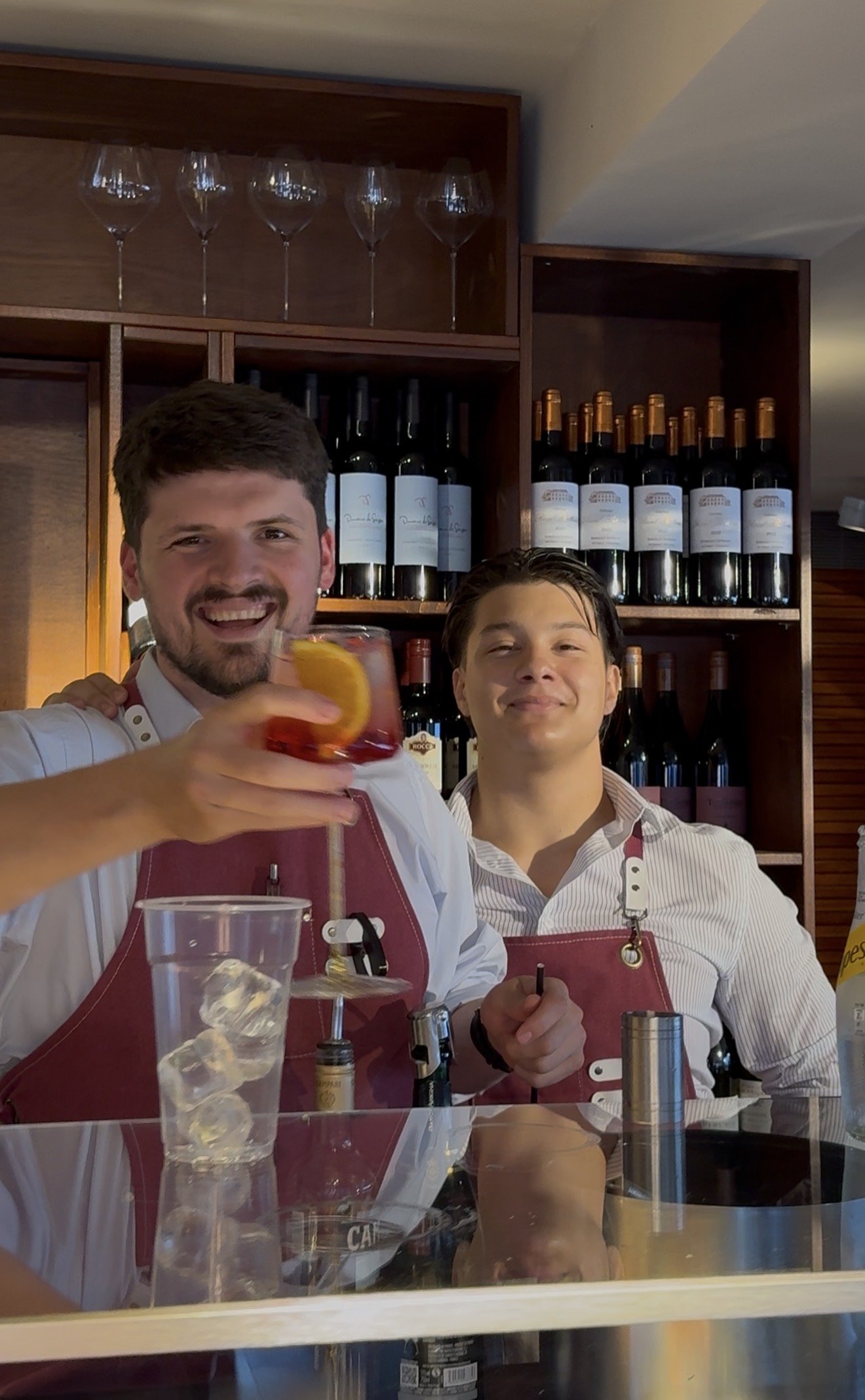 Two smiling men wearing aprons behind a bar, one holding a drink garnished with an orange slice, with wine bottles and glasses on shelves in the background.