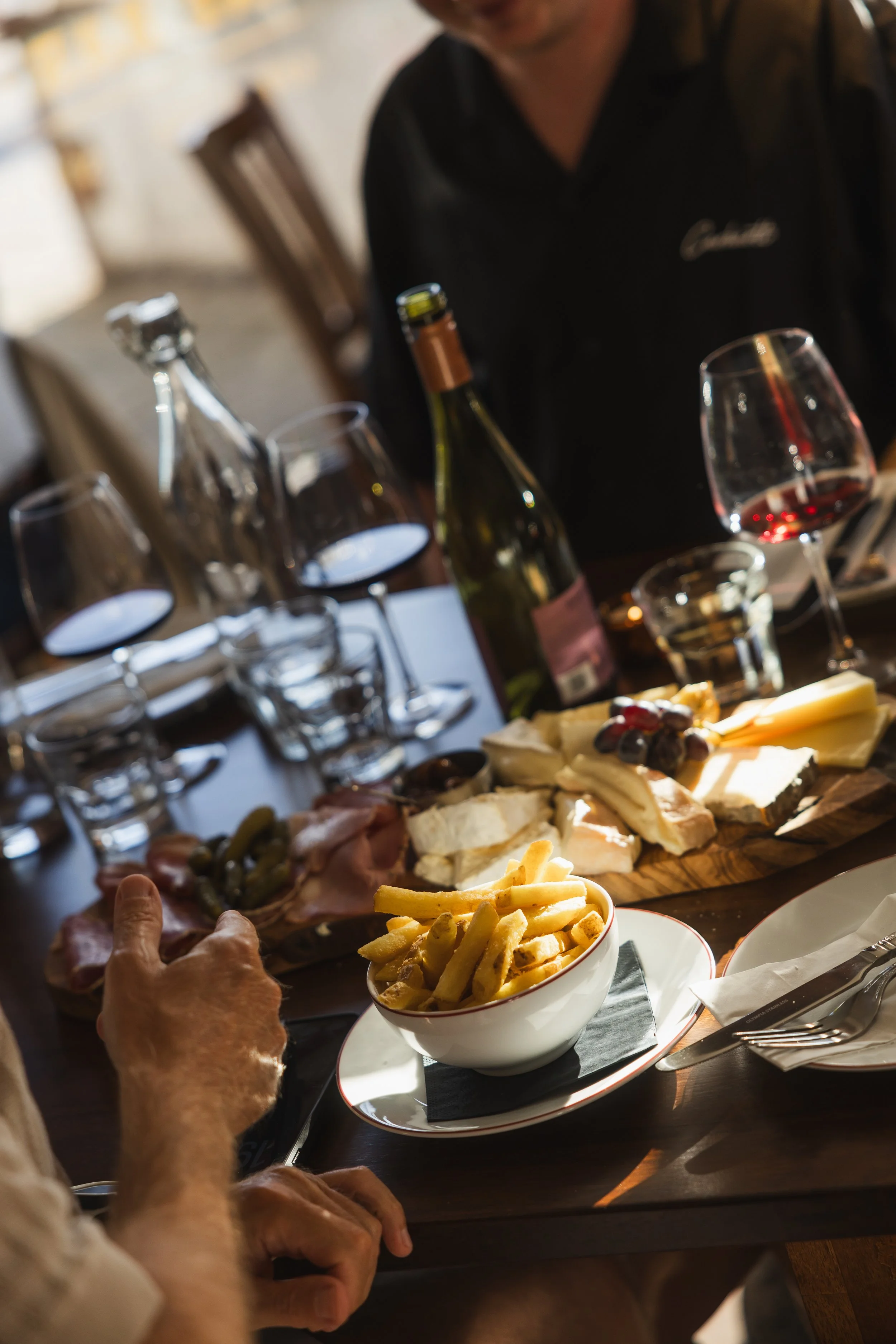 A person with a light-colored shirt is reaching for a bowl of French fries on a table at a restaurant or dining setting, with a cheese and meat platter, wine glasses, bottles, and a glass of red wine visible in the background.