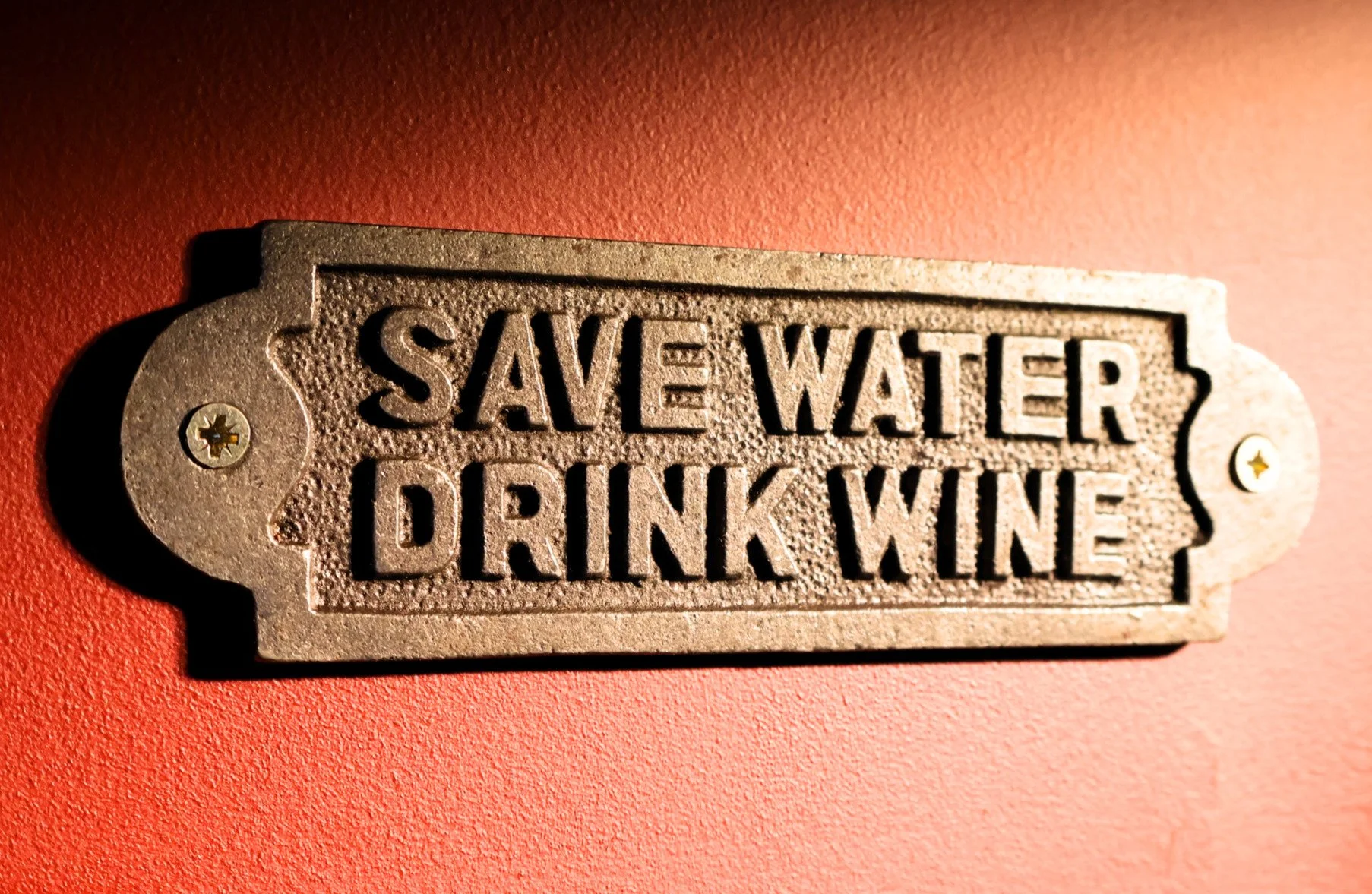Metal sign with the text "Save Water Drink Wine" mounted on a red wall.