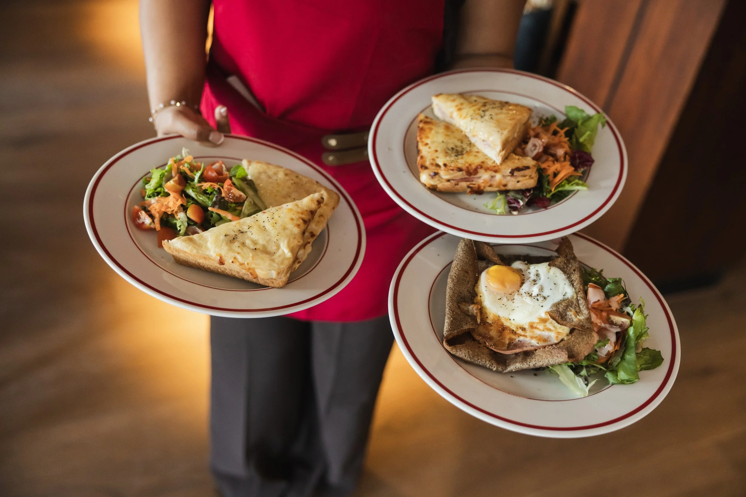 Person in red dress holding three plates with different types of food, including salad, quiche, and a dish with a fried egg on top.