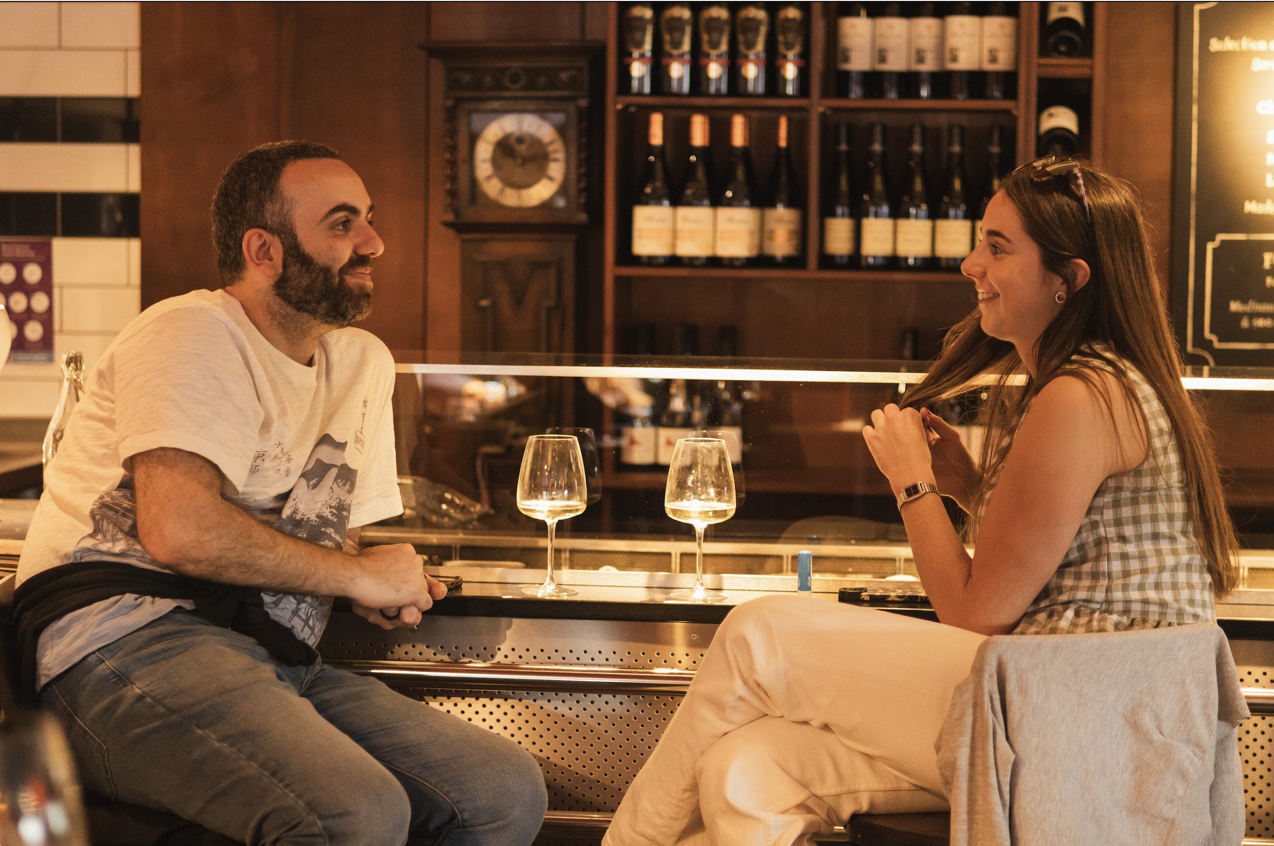 A man and a woman sitting across from each other at a bar, smiling and engaging in a conversation. There are two glasses of white wine on the bar, and the background shows shelves of wine bottles and a clock.