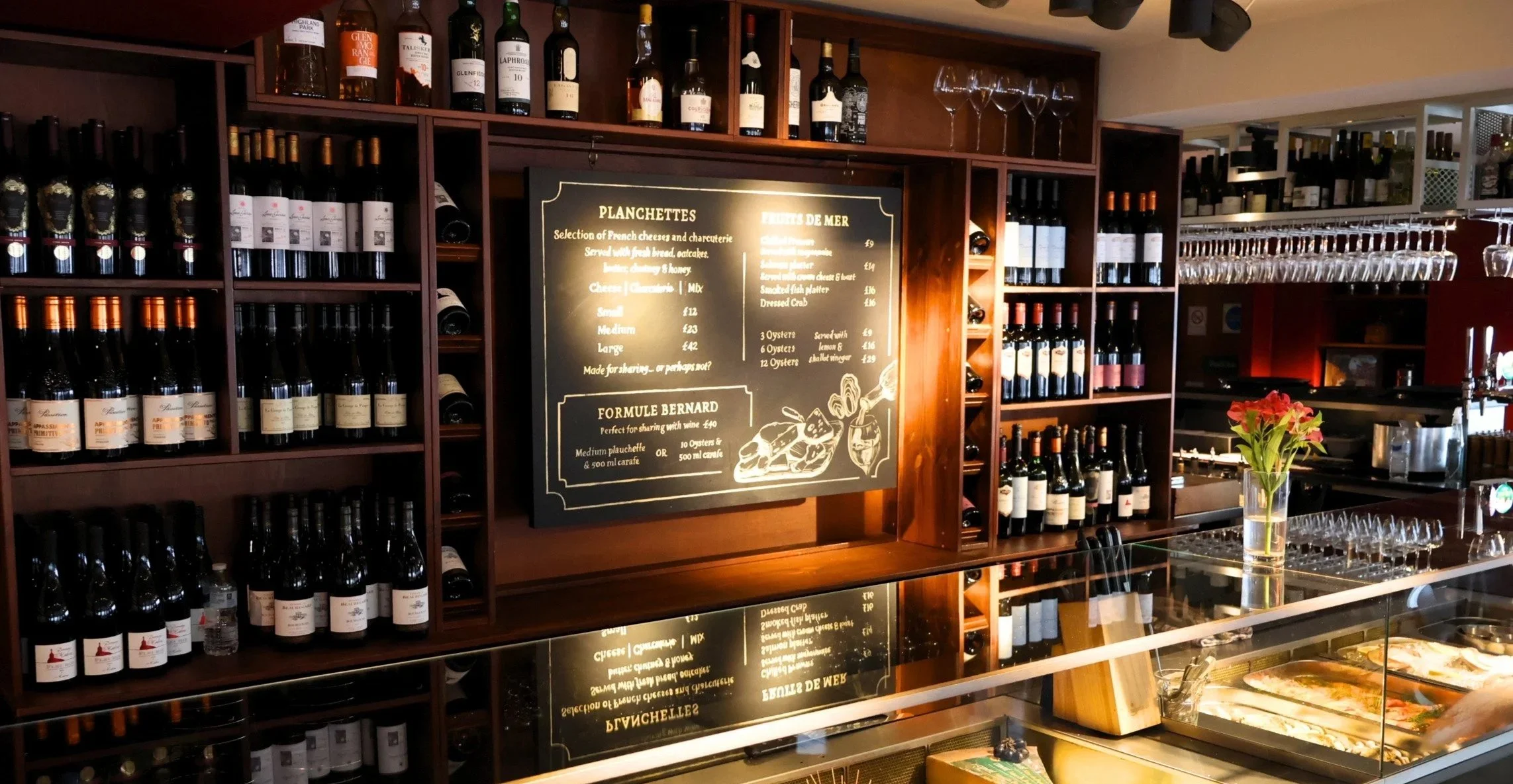 Image of a bar with wooden shelves filled with wine bottles, a chalkboard menu listing French cheeses and seafood dishes, and a bar counter with glasses and a flower vase.