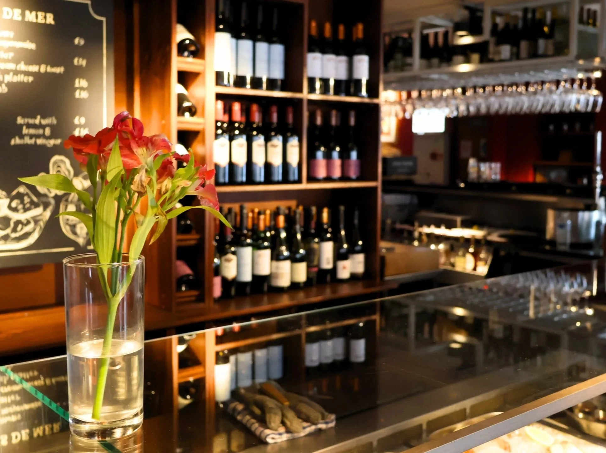 A bouquet of pink and red flowers in a tall glass vase on a restaurant counter with wine bottles and a bar in the background.