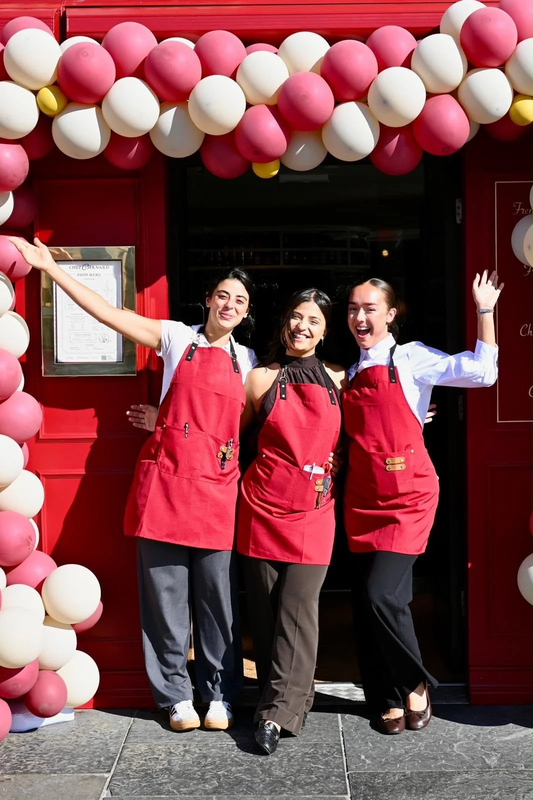 Three women in red aprons stand in front of a restaurant with a red doorway and pink, white, and yellow balloons decorating the entrance. They are smiling and posing, with two of them raising their arms.