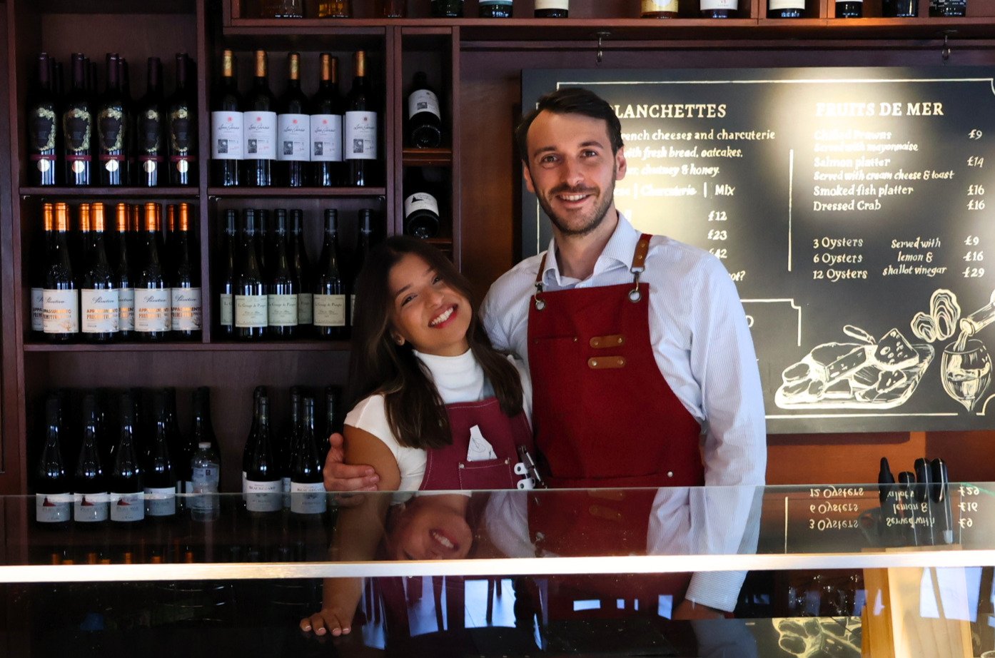 A man and a woman are smiling and standing together behind a glass counter in a wine shop or bar. The man wears a white shirt and red apron, while the woman has long dark hair, a white top, and a red apron. Behind them are shelves filled with wine bottles, and a blackboard with a menu of cheeses and seafood dishes written in chalk.
