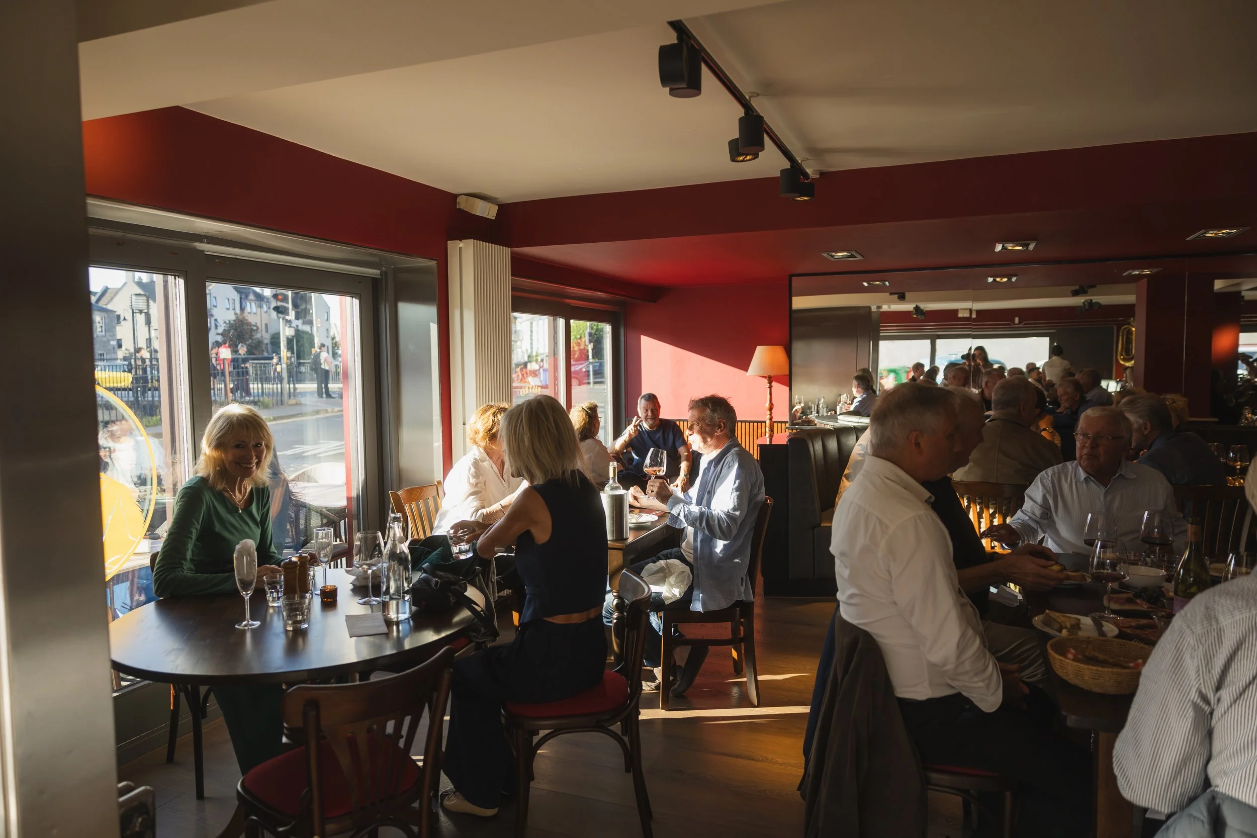 A lively restaurant scene with people enjoying their meals and conversations near large windows with sunlight streaming in.
