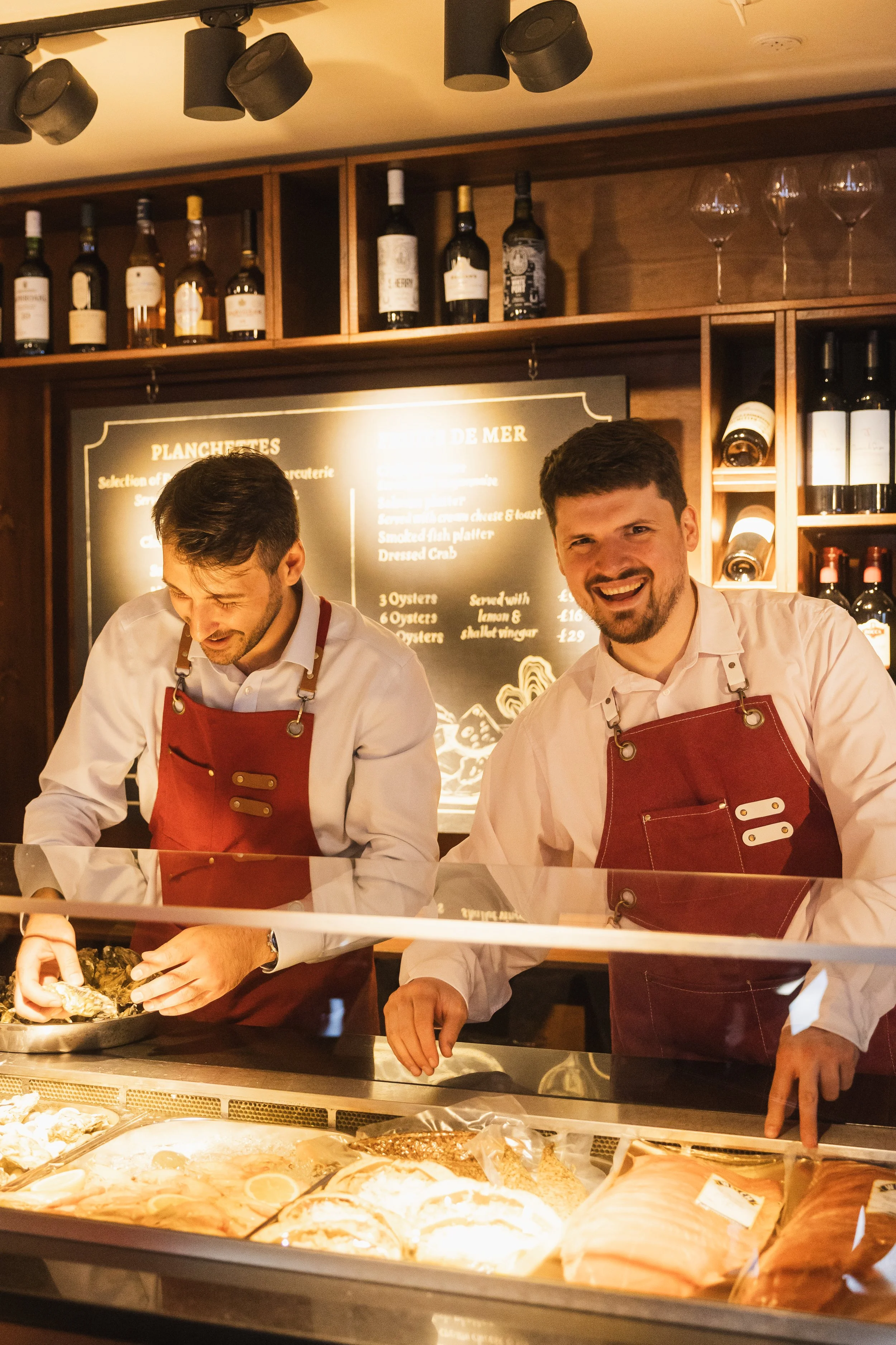 Two male chefs wearing white shirts and red aprons working at a seafood counter in a restaurant or market, with various bottles of wine and glasses on shelves behind them and a blackboard with menu items in the background.