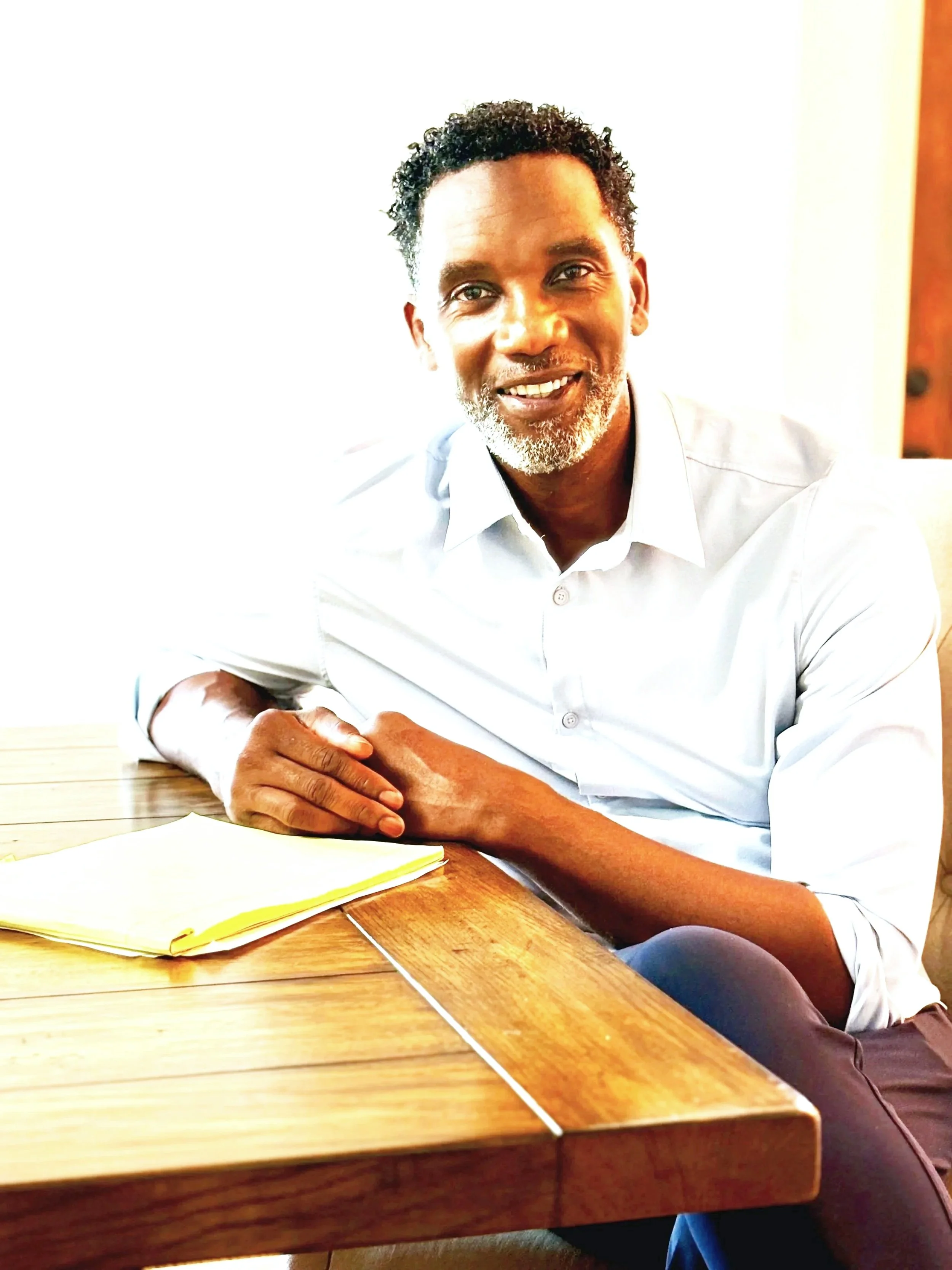A smiling Black man with curly hair and a beard, wearing a white shirt, sitting at a wooden table with his arms crossed, next to a person whose arms are partly visible.