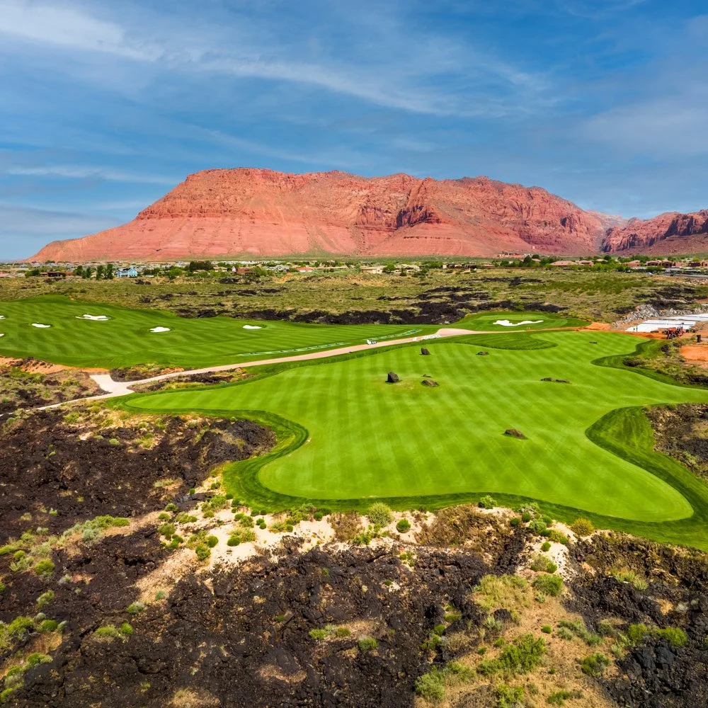 A golf course with lush green fairways and sand traps, set against a backdrop of red rock formations and a blue sky.