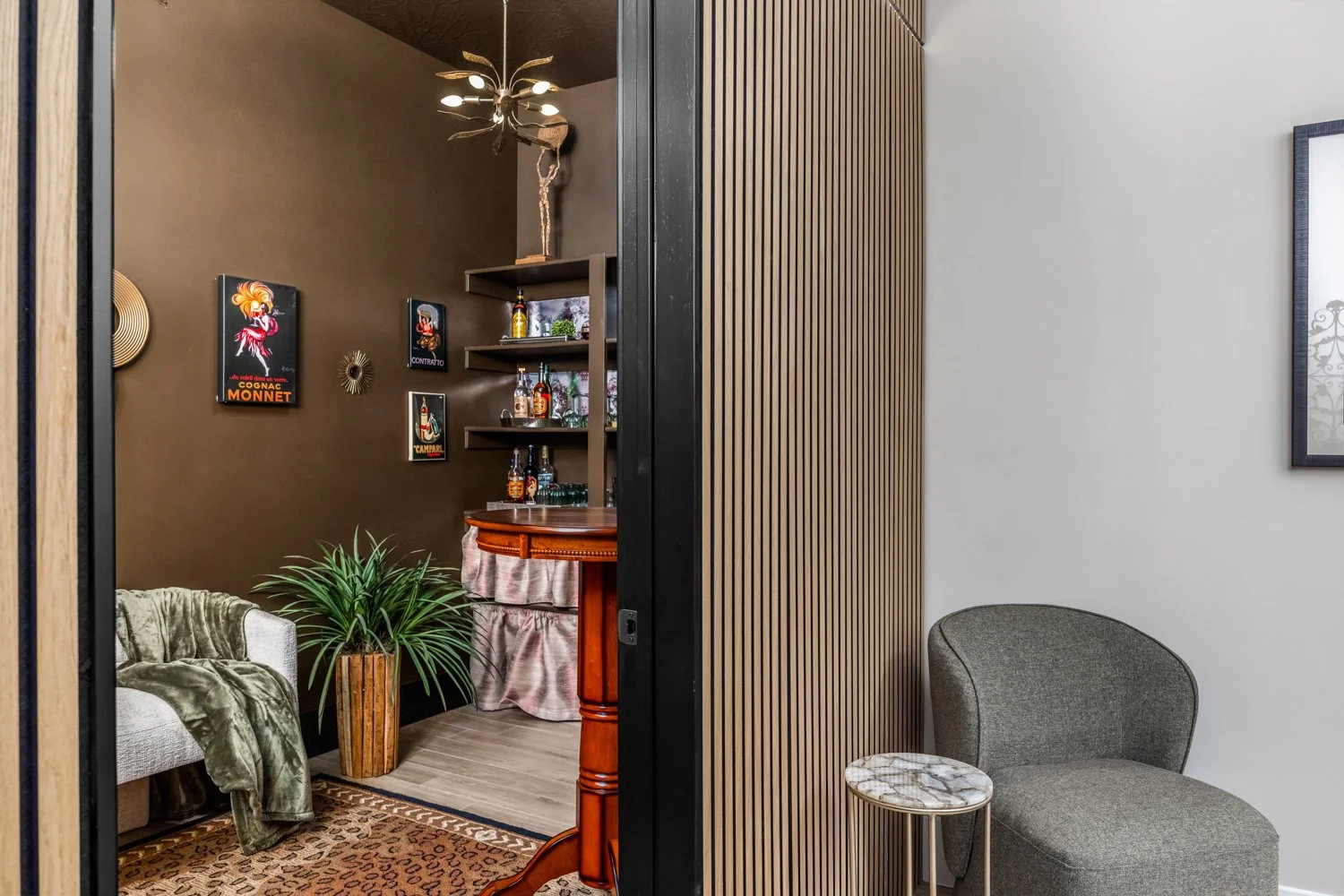 A cozy living room with a gray armchair, a small marble-top side table, and a large indoor plant. The room is divided by a wooden slatted partition, with a dark accent wall decorated with vintage posters and a gold wall sculpture. A chandelier hangs 