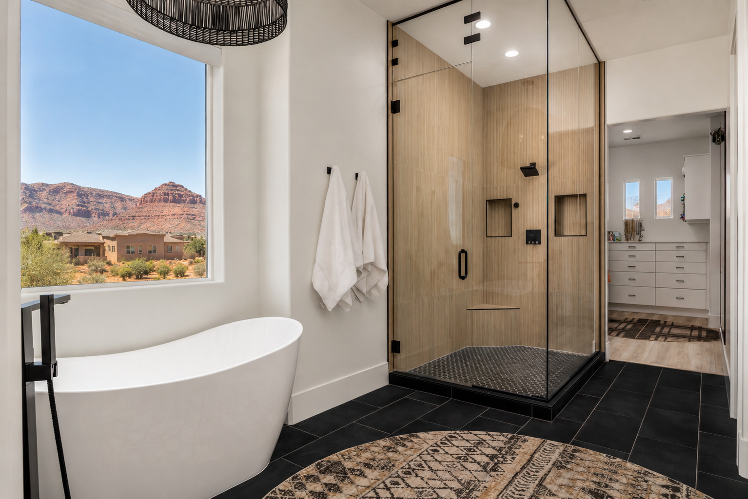 Modern bathroom with a black tiled floor, freestanding white bathtub, glass-enclosed shower with wood walls, white towels hanging, and a window showing a desert landscape with mountains.
