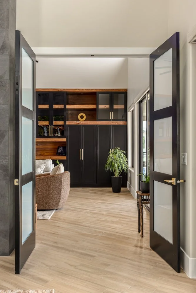 Open black-framed glass doors leading into a living room with wooden floors, black cabinets, and potted plants.