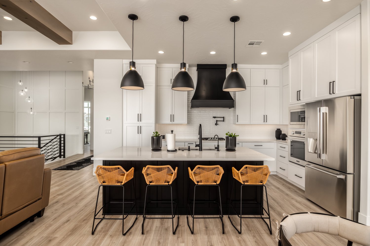 Modern kitchen with white cabinets, a large island with black base, four black pendant lights, and four orange bar stools, stainless steel refrigerator, and microwave, with hardwood flooring.