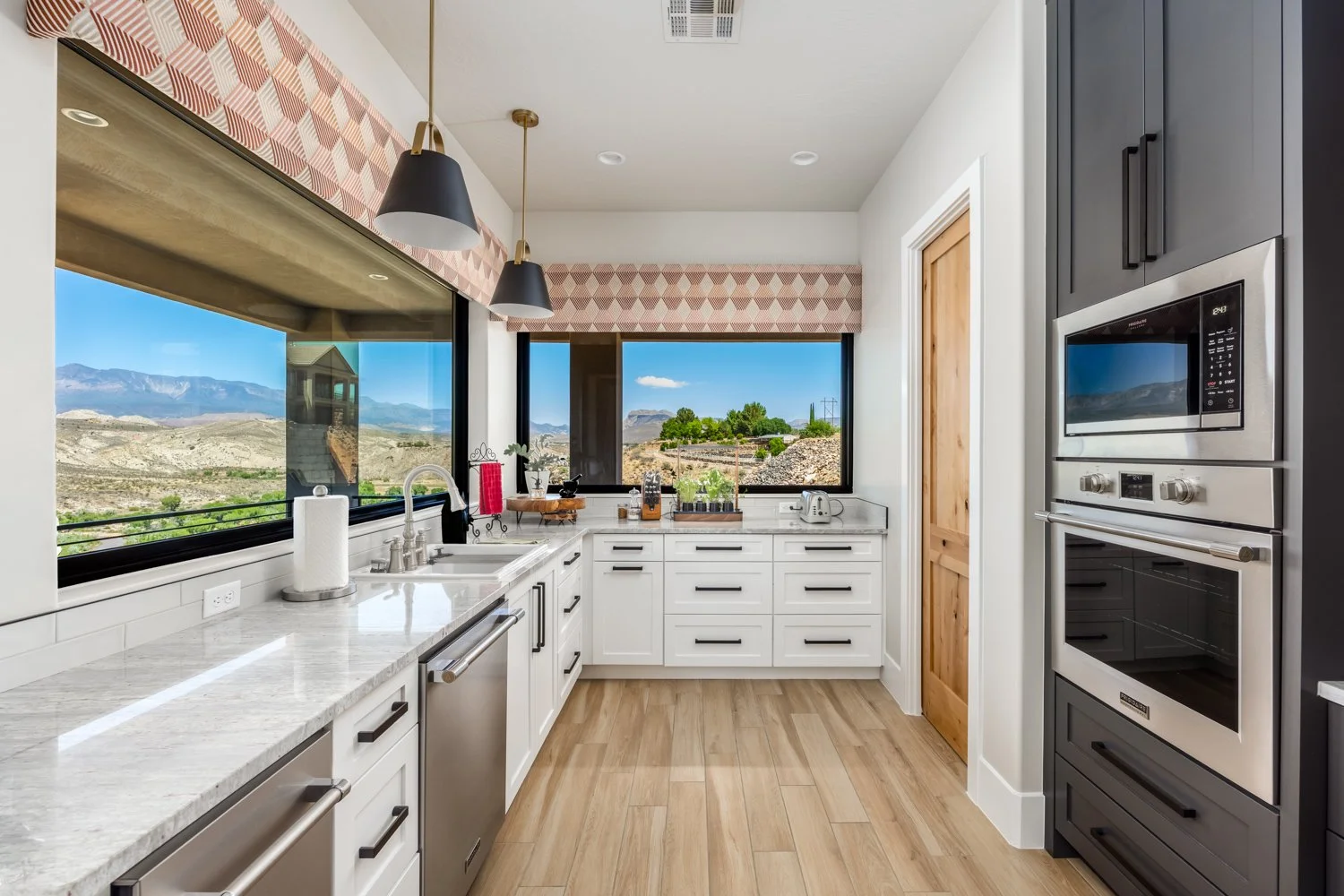Modern kitchen with white cabinets, black accents, wood flooring, large windows showing mountain and desert landscape, and stainless steel appliances.