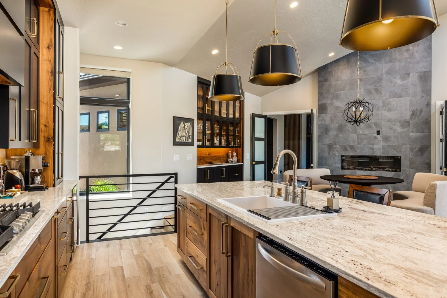 Modern kitchen with wooden cabinets, a large marble island, black pendant lights, a gray tiled fireplace wall, and a sitting area with armchairs and a small round table.