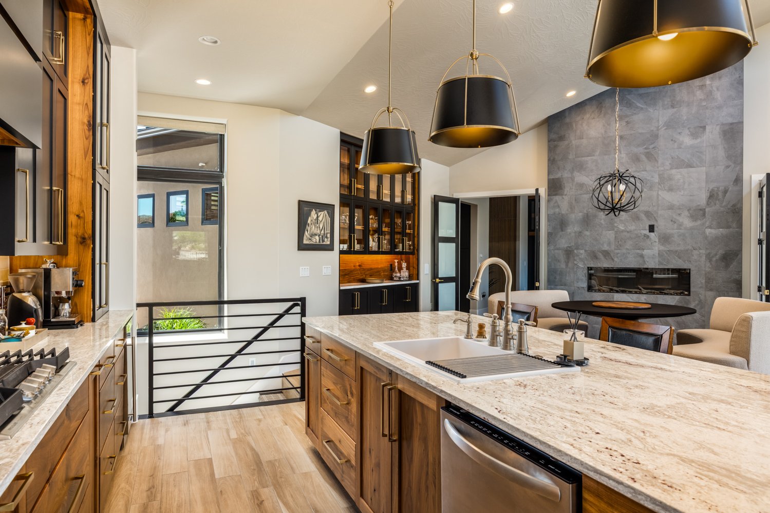 Modern kitchen with wooden cabinets, marble countertop island, black and gold pendant lights, gray stone fireplace, and a seating area with beige armchairs and a black round table.