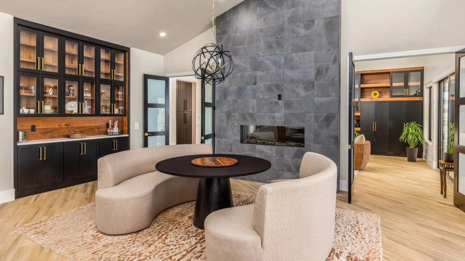 Modern living room with a round black table, curved beige sofa, and a black and wood cabinet against the wall; a gray stone fireplace wall is in the background, with a geometric black chandelier hanging from the ceiling.