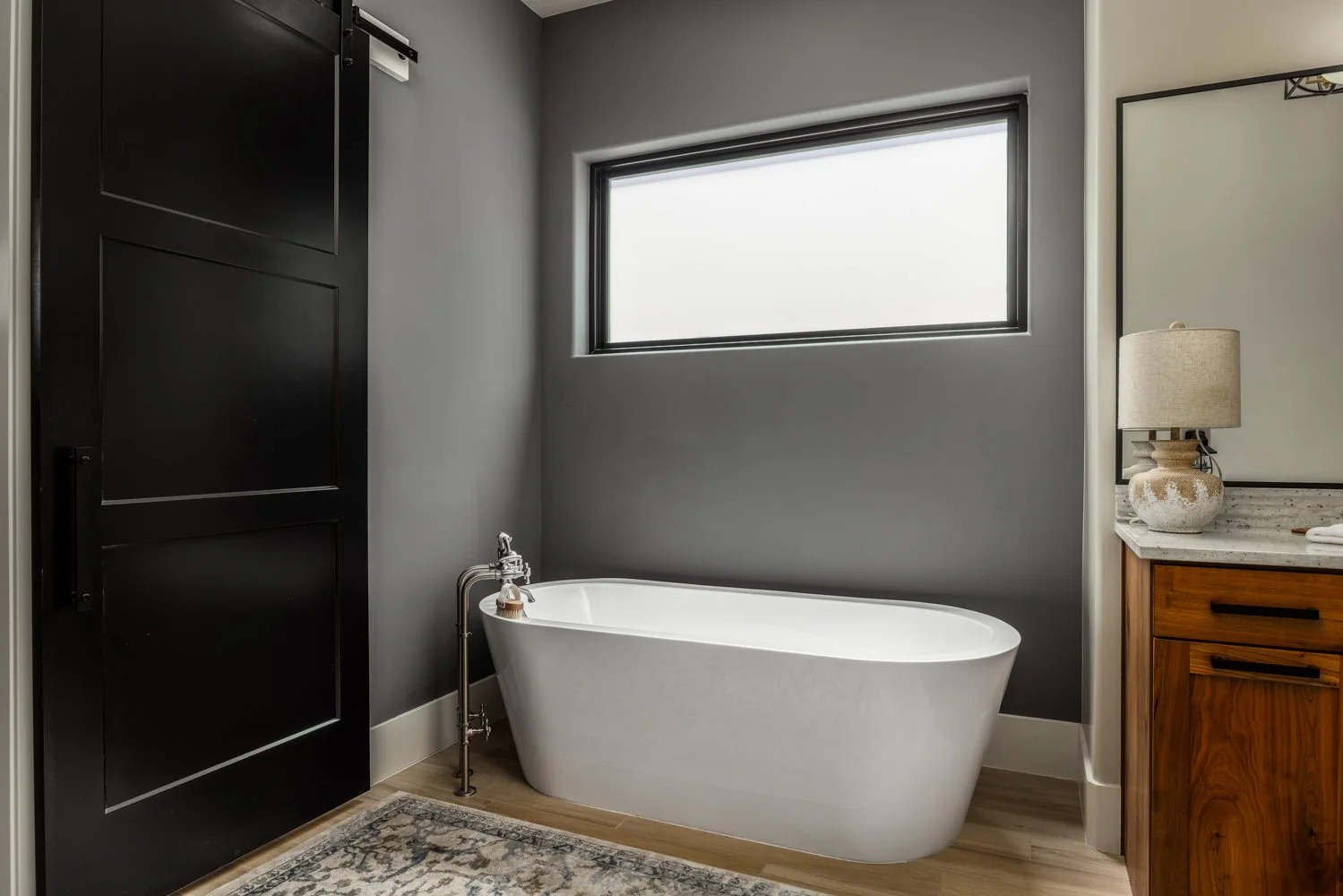 Modern bathroom with a white freestanding bathtub, a gray wall, a large window, a wooden vanity with a lamp, and a decorative rug.