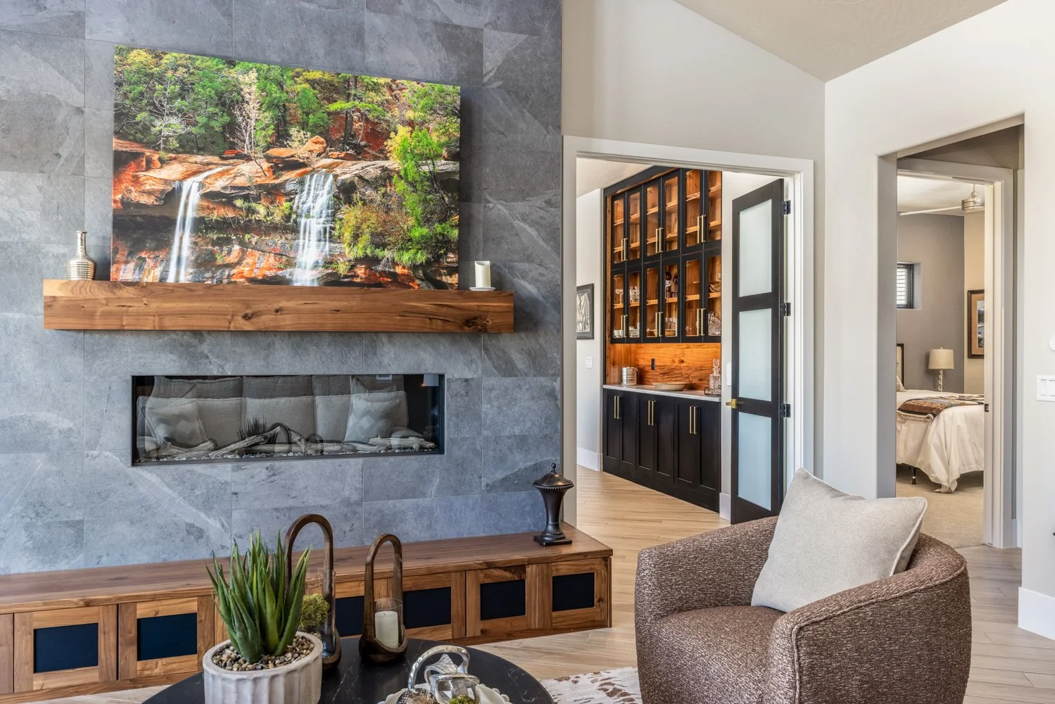 Modern living room with gray tiled fireplace, framed waterfall photograph above mantel, and wooden console with decorative items. Adjacent to a cozy armchair and a view into a kitchen and bedroom.