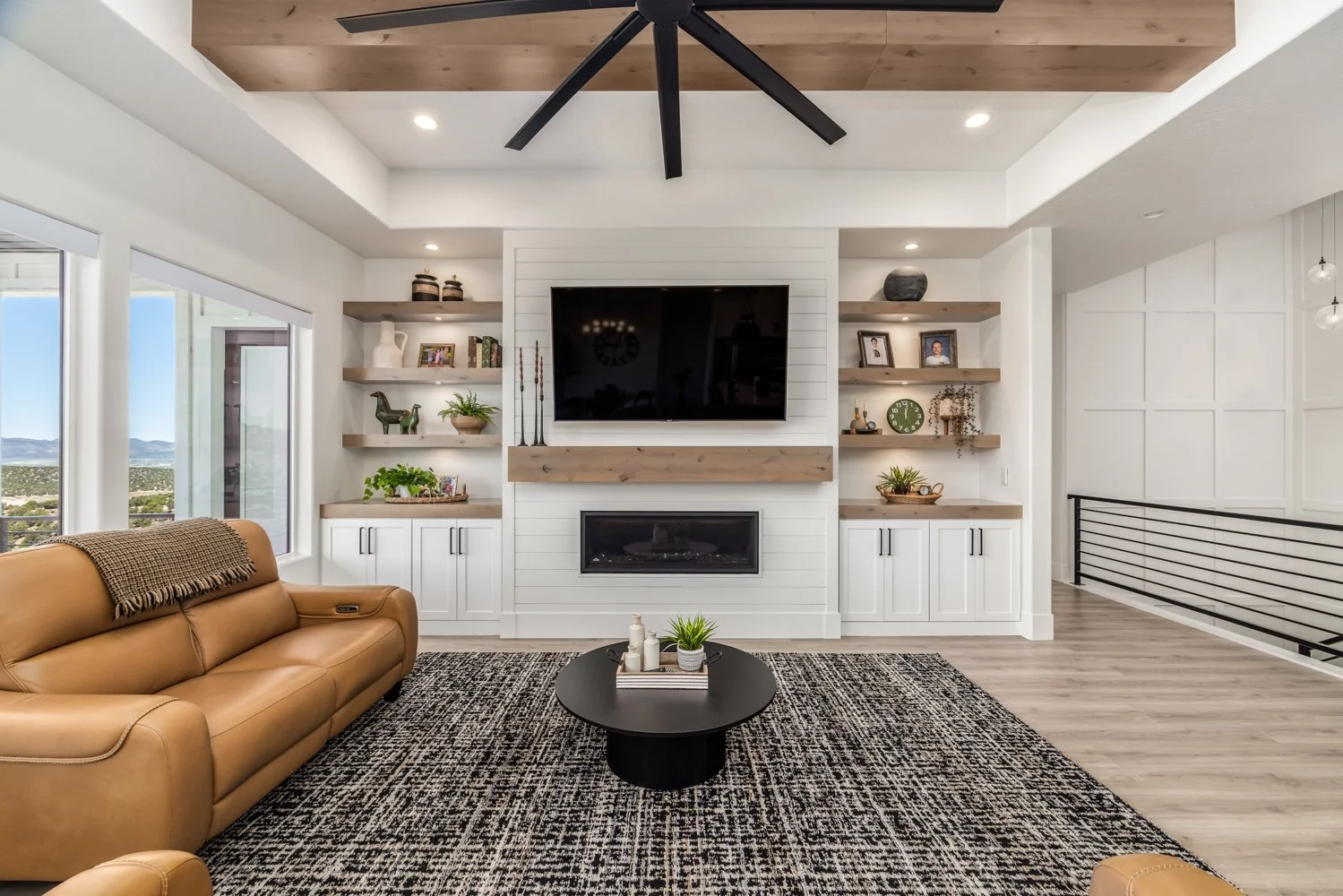 Living room with a tan leather couch, a black coffee table with a plant and decorative items, large windows showing a landscape, built-in white shelves with decorative items, a wall-mounted TV, a fireplace, wood laminate flooring, and a ceiling fan.
