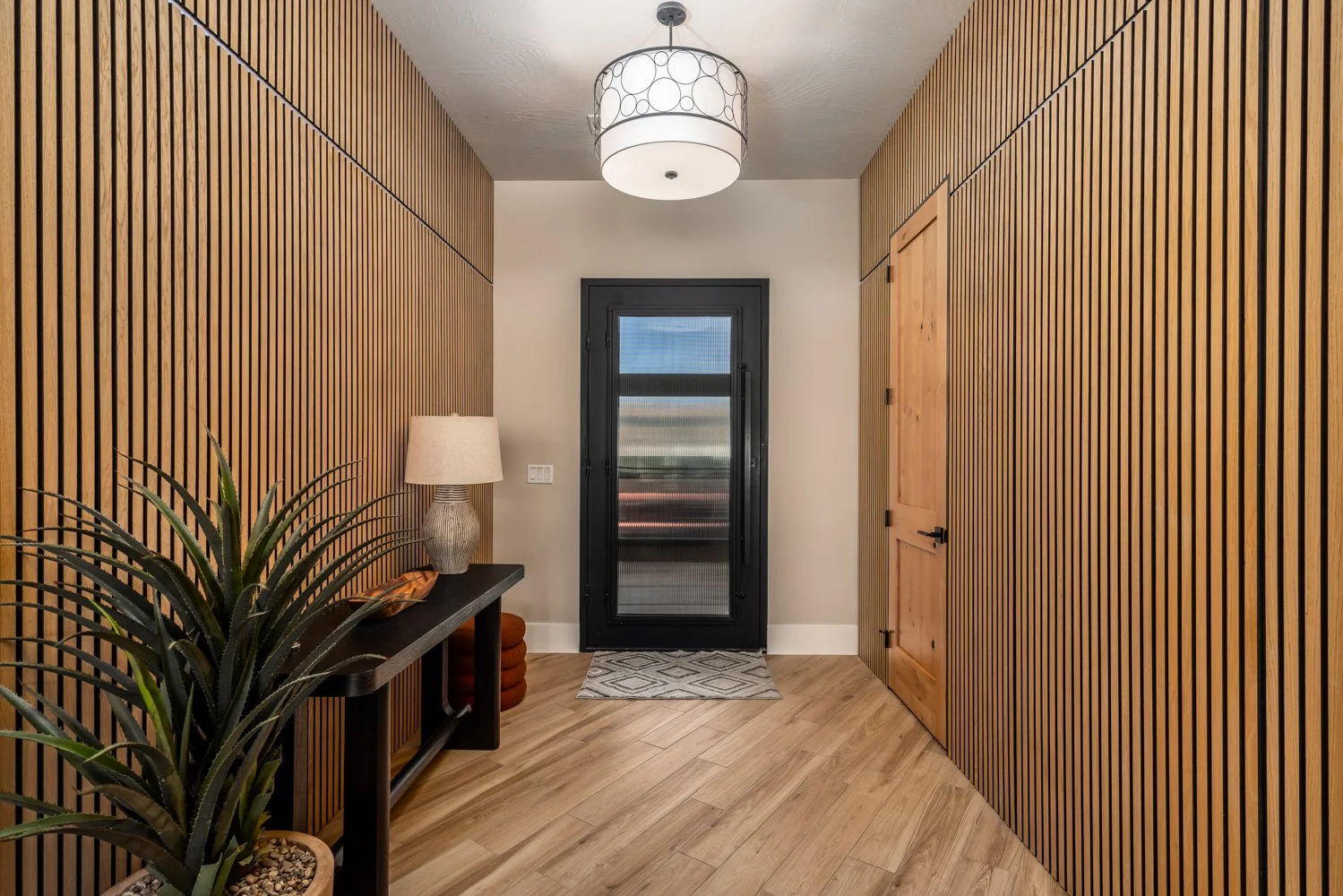Entryway with wooden slat walls, a black door with a glass panel, a wooden door, a console table with a lamp and decorative items, a potted plant, and a ceiling light fixture.