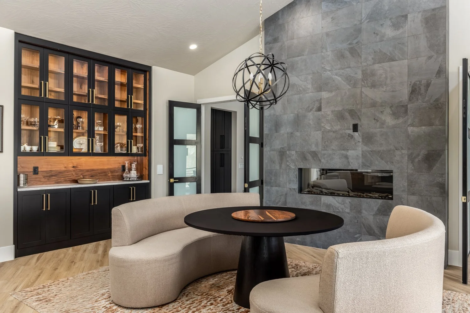 Modern living room with a curved beige sofa, a round black table with a wooden tray, a beige armchair, a textured gray wall with a built-in fireplace, black and wood cabinetry, frosted glass doors, and a geometric black chandelier.