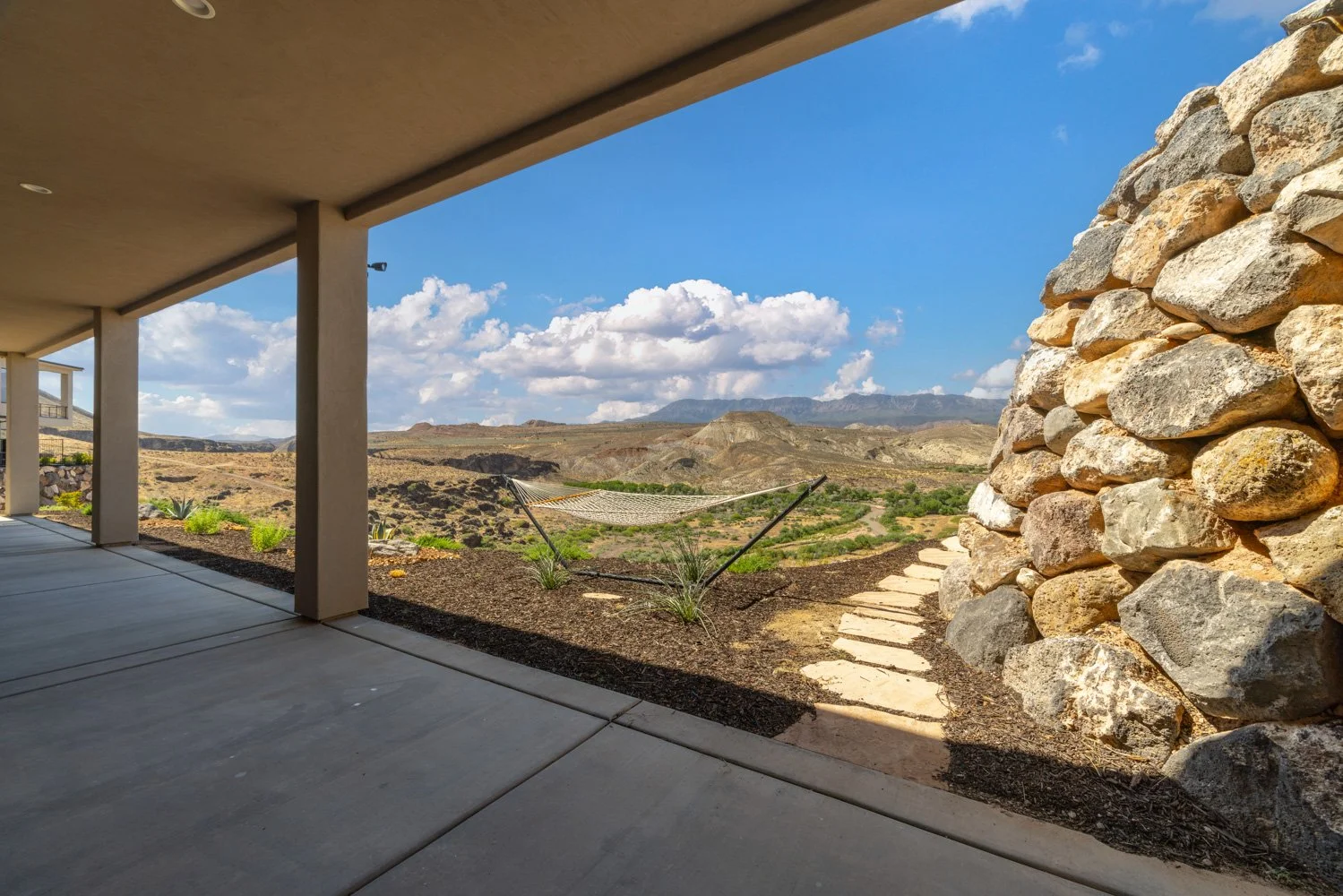 A covered patio overlooking a desert landscape with mountains in the distance, featuring a hammock between two plants and a stone pathway.