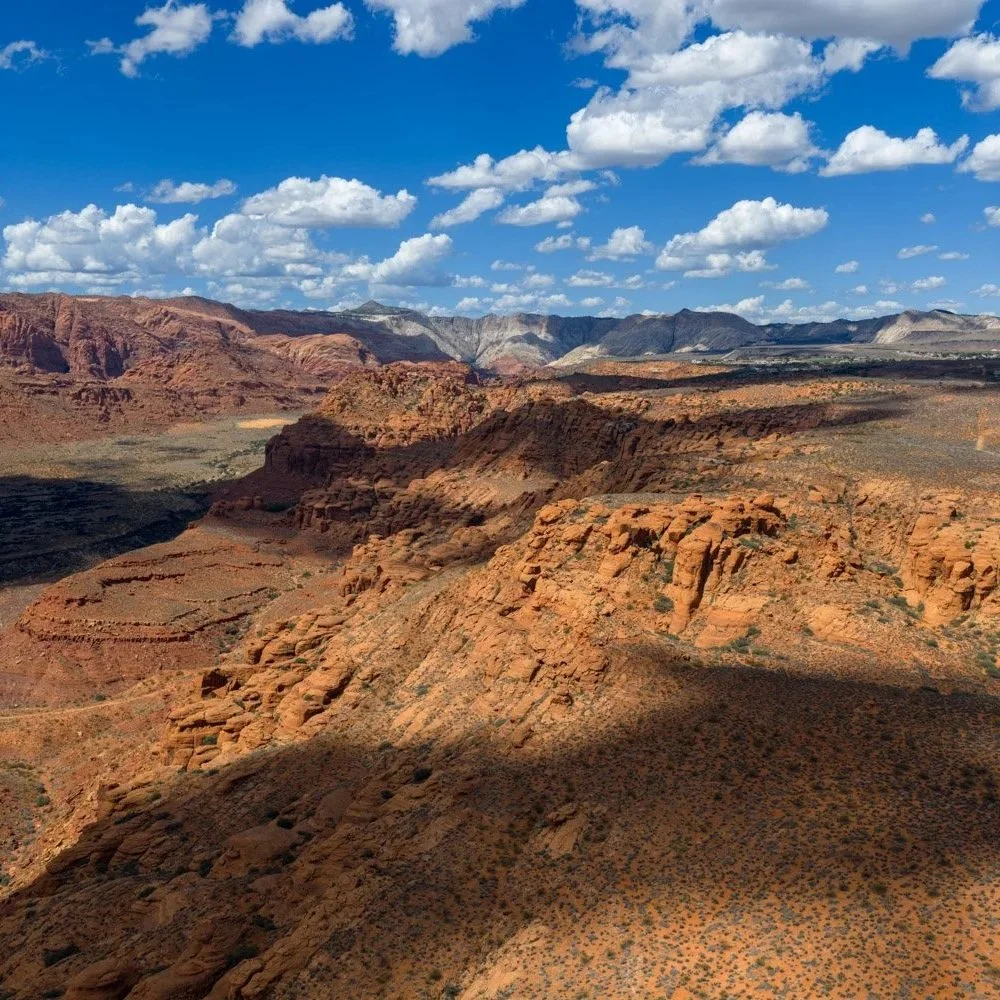 Scenic view of a desert landscape with red rock formations, distant mountains, and a partly cloudy blue sky.