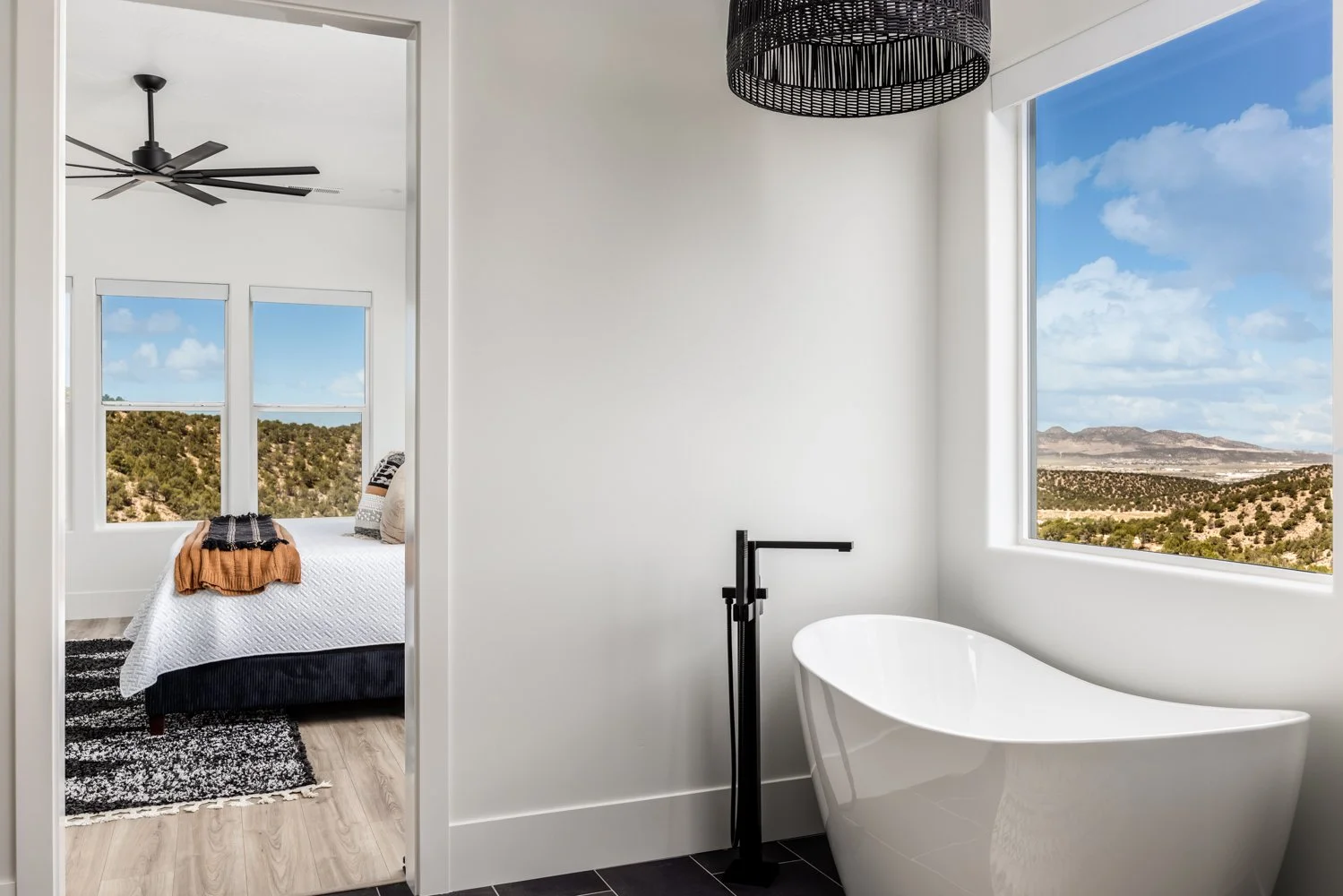 Bathroom with white bathtub under large window showing desert landscape, bedroom visible through door with bed, black ceiling fan, and part of black and white rug.