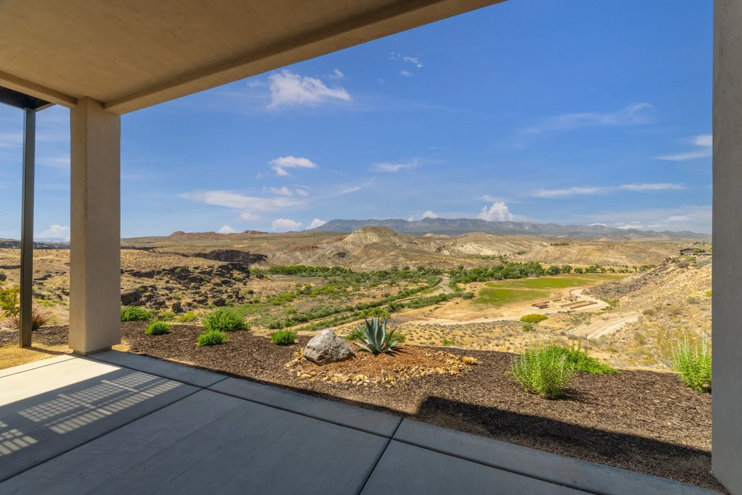View from a covered patio looking out over a desert landscape with rolling hills, sparse vegetation, and a clear blue sky.