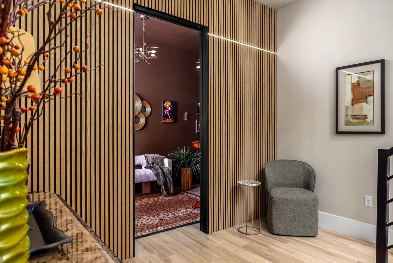 Interior of a modern living space with a wooden slat wall, a gray chair, a small round side table, and artwork on the wall. A doorway leads to a room with a purple wall, a sofa, and decorative art.