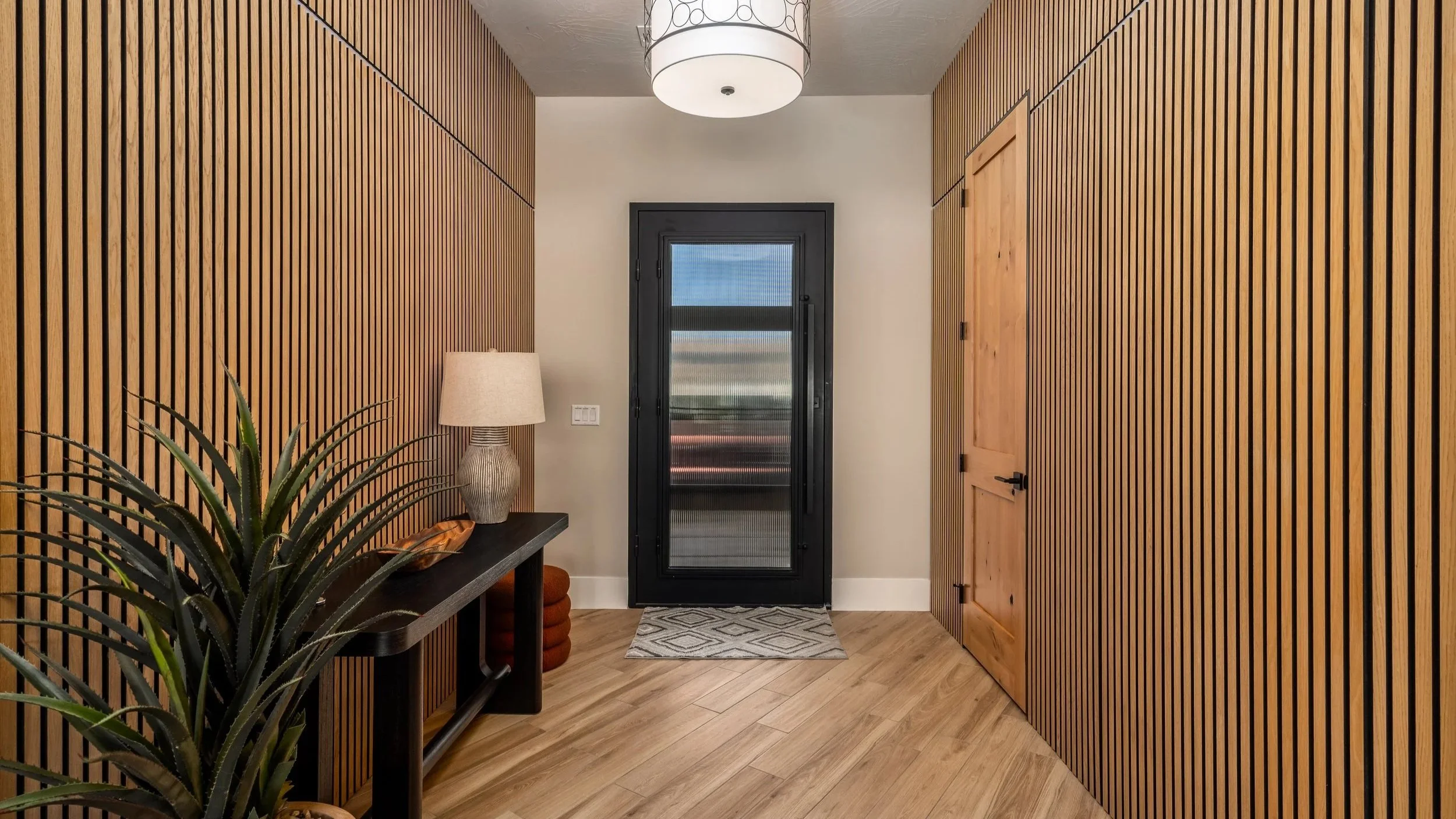 Modern entryway with light wood slat walls, a black front door with glass, a round ceiling light, a side table with a lamp, and a wooden door on the right.